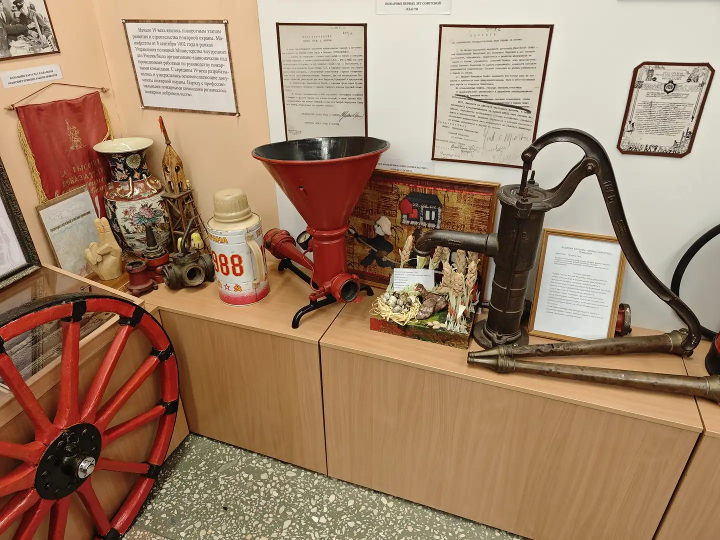 展台上摆放着各种老式消防设备，包括红色的轮子、金属漏斗、手摇泵和灭火器等。墙上挂有几份文件和说明牌，介绍这些设备的历史和用途。展台左侧有一个红色的陶瓷罐，右侧则放置了一些小型模型和工具。