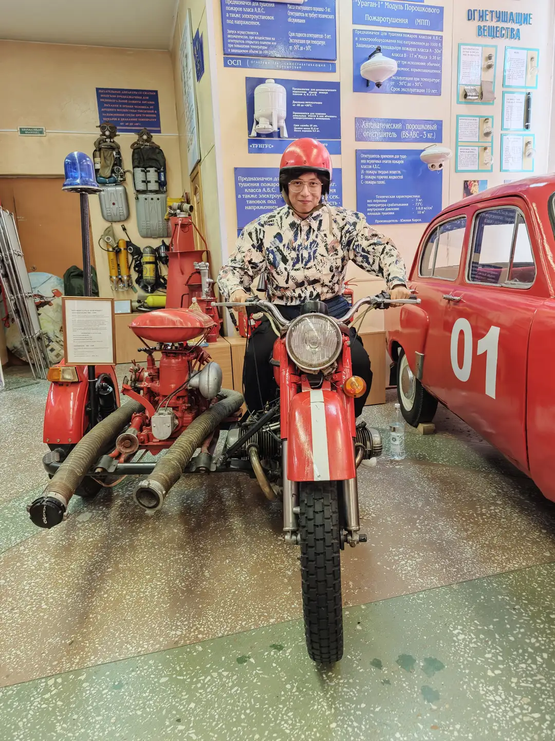 一个人戴着红色头盔，坐在一辆红色的消防摩托车旁，摩托车后部装有消防设备。旁边停着一辆红色的老式汽车，车牌号为01-85АМО。背景墙上挂满了消防器材和说明牌，展示各种消防设备和知识。