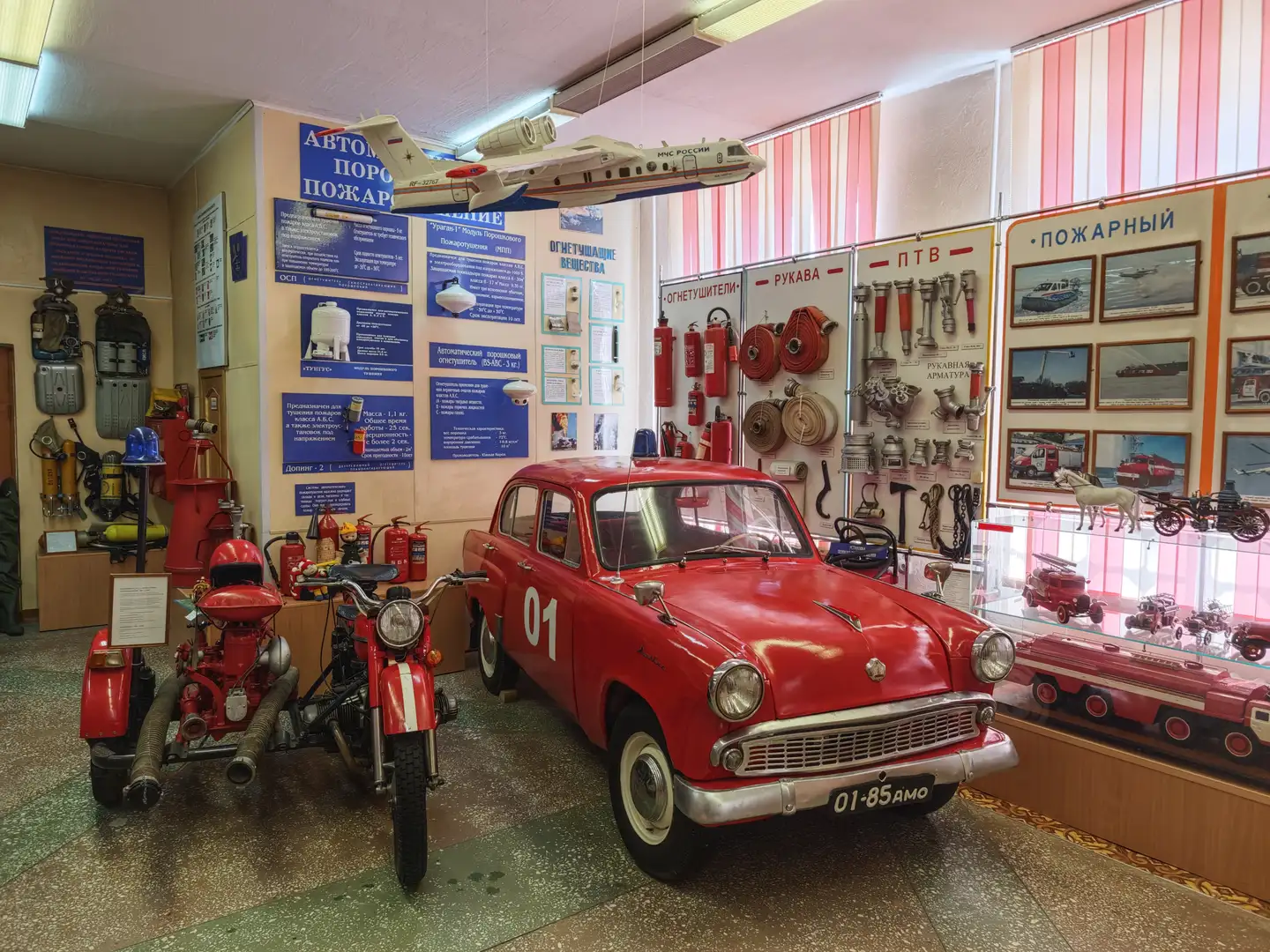 展厅内陈列着一辆红色的消防摩托车和一辆红色的复古汽车，周围墙上挂满了消防器材和说明牌。天花板上悬挂着一架白色飞机模型，右侧展柜中展示着各种消防车辆模型。整个空间布置得井然有序，充满历史感。