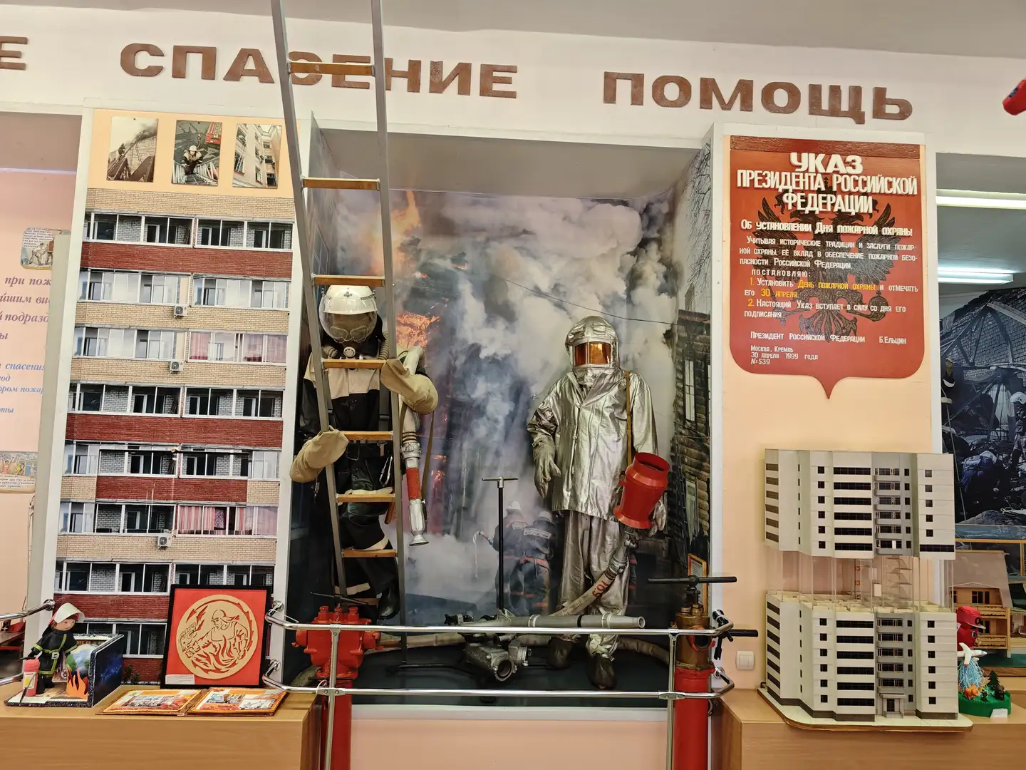 展览区中央有两个身穿防护服的假人模型，一个攀爬梯子，另一个手持消防水枪。背景是一幅燃烧的大楼画面，上方写着“救援”和“帮助”。右侧展示了一块总统令公告，左侧有建筑物模型和消防标志，整体呈现火灾救援场景。