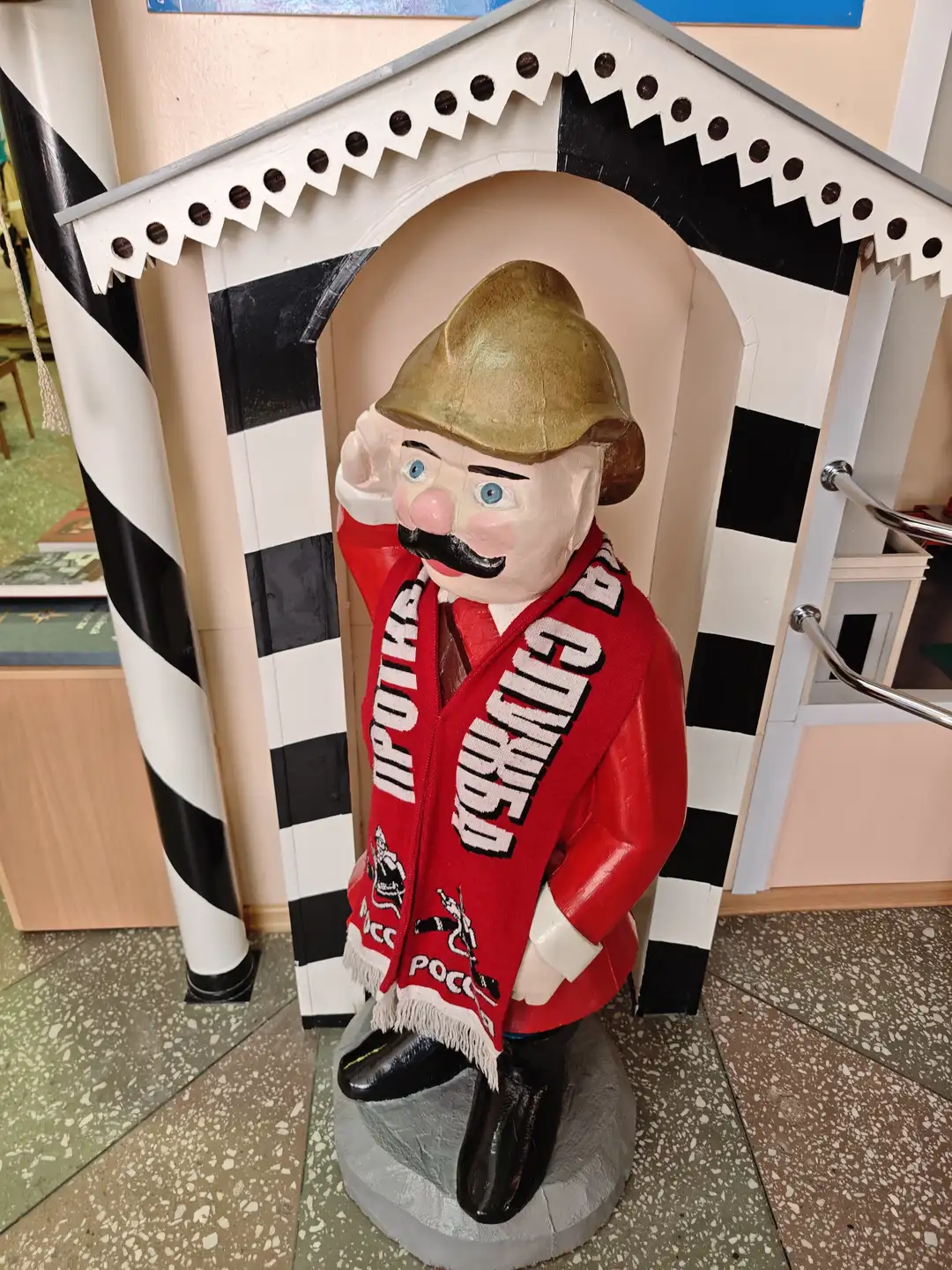 一个穿着红色外套、戴着金色帽子的卡通人物雕像站在黑白相间的建筑前，雕像的外套上写有“ПОЖАРНАЯ ОХРАНА”字样。雕像右手敬礼，左手插在口袋里，脸上有浓密的黑色胡子和蓝色眼睛。背景是带有条纹图案的墙壁和柱子，地面为斑点状瓷砖。