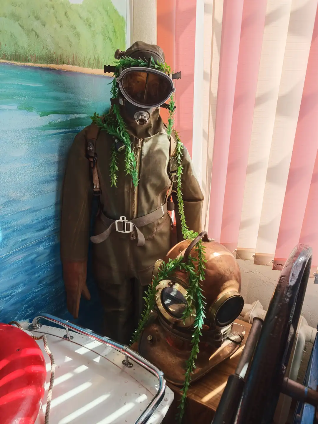 一名穿着深色潜水服、戴着防毒面具的人物模型站在船边，面具和衣服上缠绕着绿色藤蔓。旁边放置着一个铜制潜水头盔，同样装饰有藤蔓。背景是一幅描绘河流和岸边风景的壁画，周围有金属栏杆和船只部件。