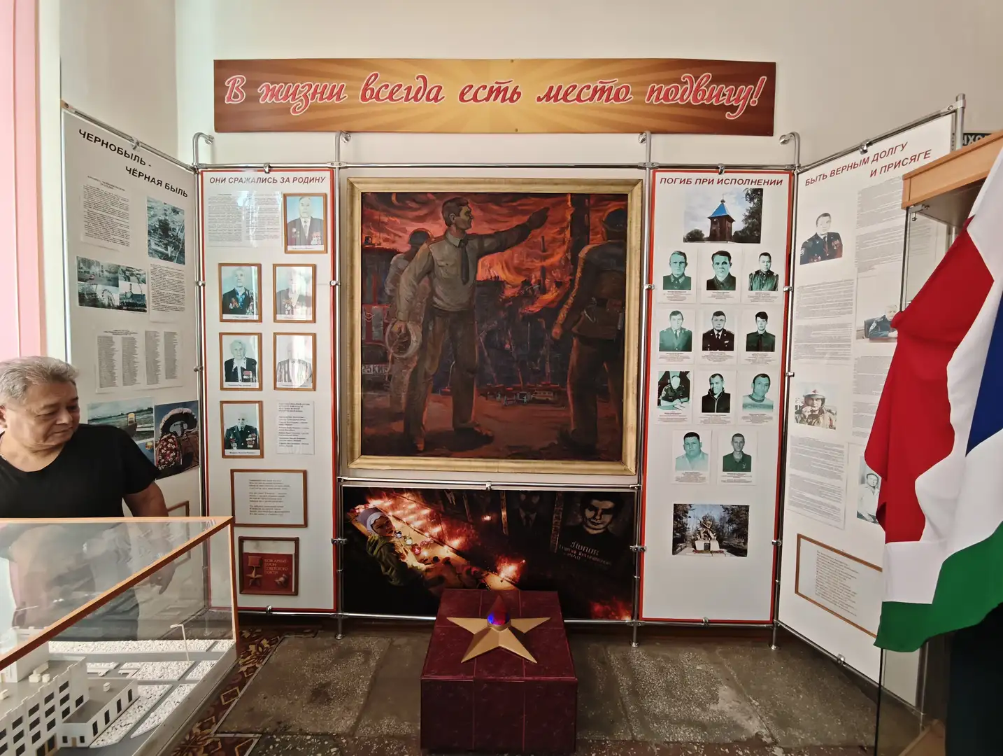 展览墙上有大幅画作和多张照片，中央画作描绘了两名男子在火场中指挥救援的场景。上方横幅写着“В жизни всегда есть место подвигу!”，两侧展板展示了人物肖像和文字介绍。前方有一个红色盒子，上面放置着一颗金色五角星，旁边有玻璃展柜和一面三色旗。