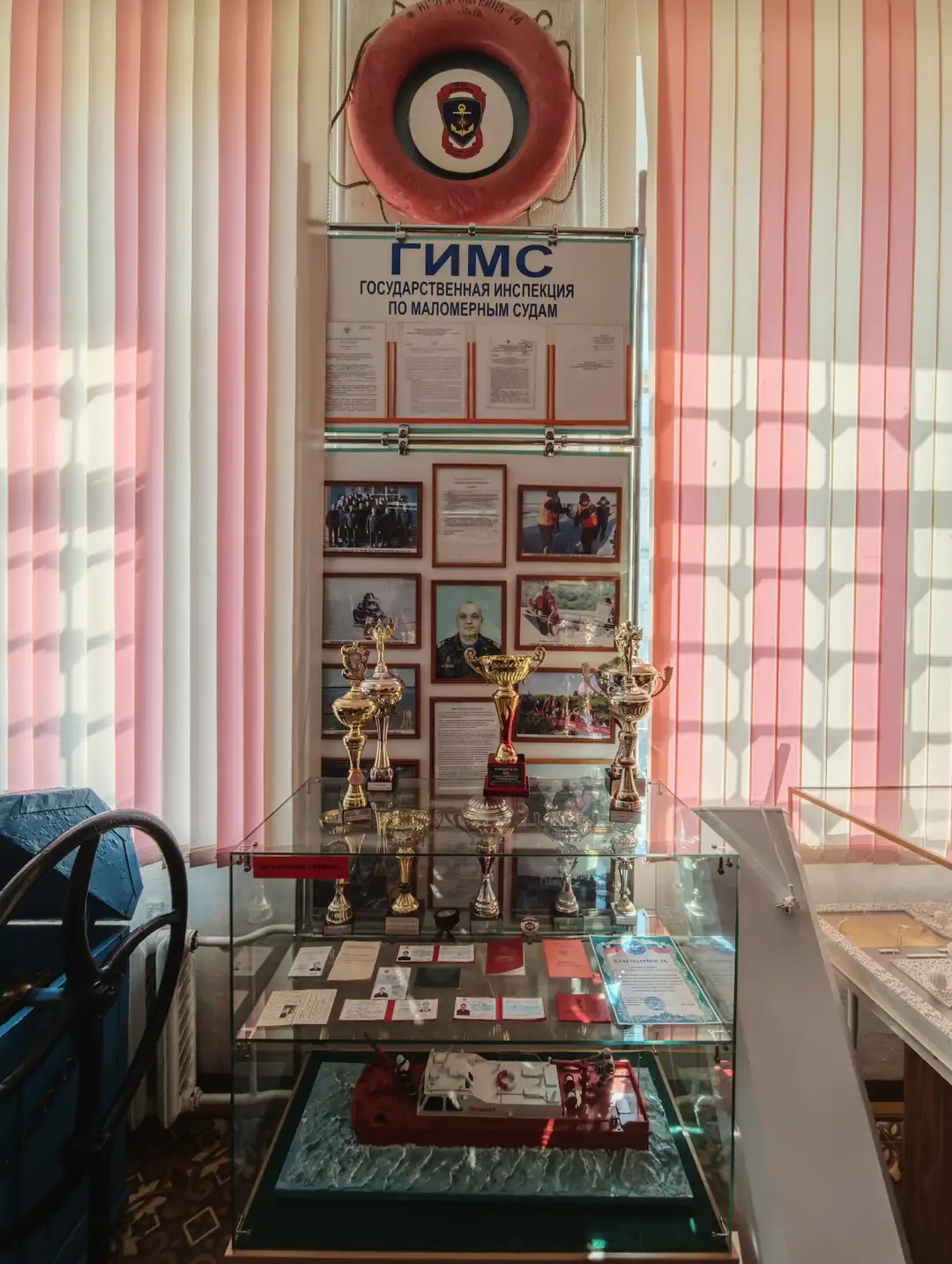 展示柜中陈列着多个奖杯和证书，上方悬挂着一个红色救生圈，救生圈中心有徽章图案。展柜后方的墙上贴有多张照片和文件，上方标有“ГИМС ГОСУДАРСТВЕННАЯ ИНСПЕКЦИЯ ПО МАЛОМЕРНЫМ СУДАМ”。展柜底部展示了一艘红色小船的模型，周围有文件和证件。