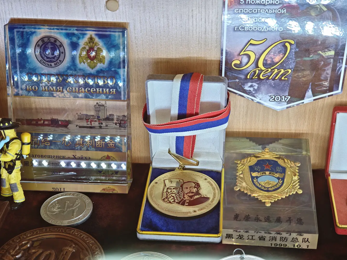 展台上摆放着一枚金色奖章，奖章上刻有两个人物肖像，系着红蓝白三色绶带。左侧是一个透明展示盒，内有消防主题的纪念品和中文标语“团结一心 其利断金”。右侧是一个金色徽章，下方有中文文字“黑龙江省消防总队 1999.10.1”，旁边还有一张庆祝50周年的海报。