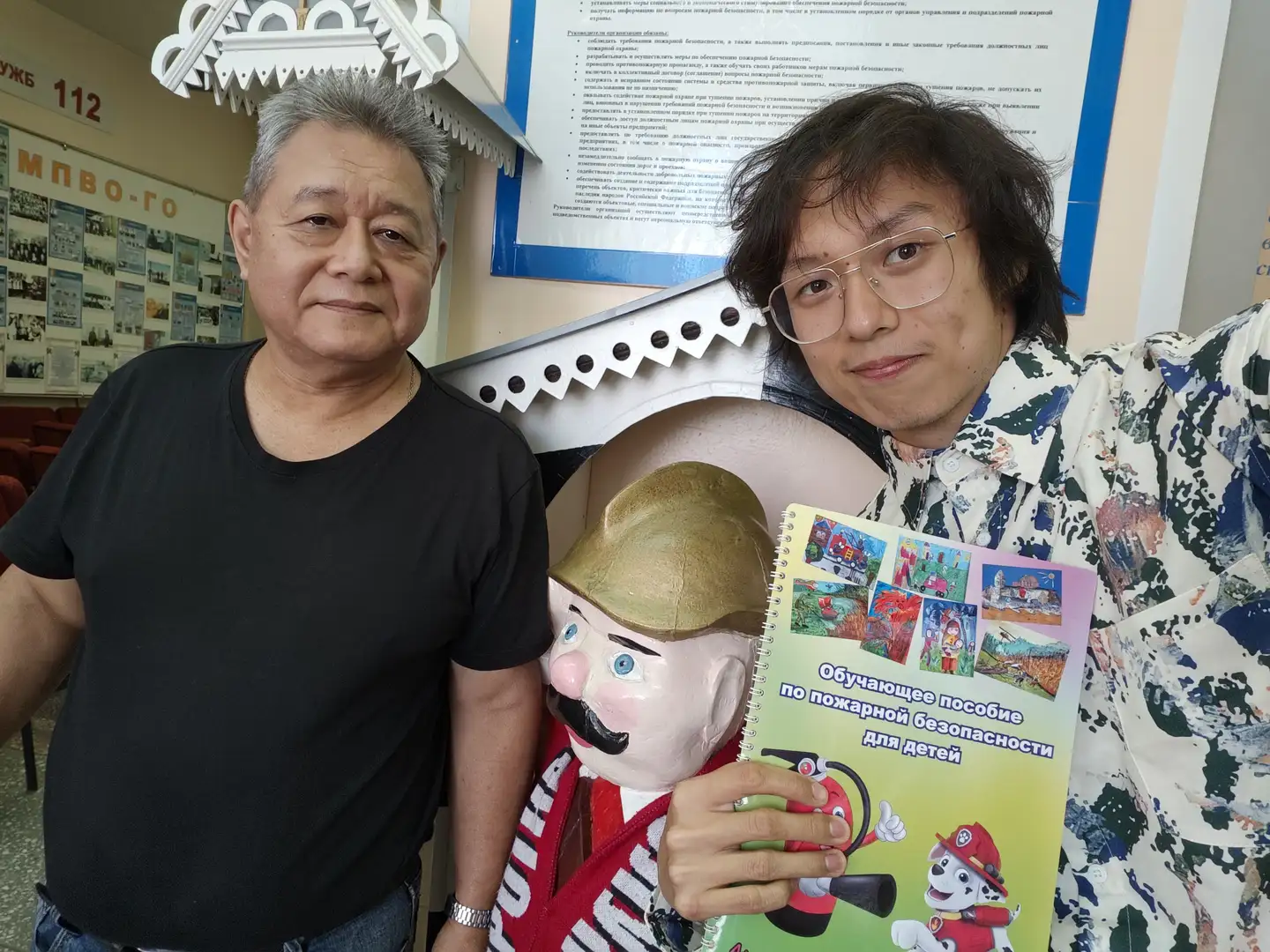 两位男士站在一个卡通消防员雕像前合影，其中一人手持一本封面印有消防狗图案的彩色儿童读物。背景墙上贴有消防知识宣传资料，上方有数字“112”和俄文标识。两人面带微笑，身后环境为室内，有装饰性屋顶结构。