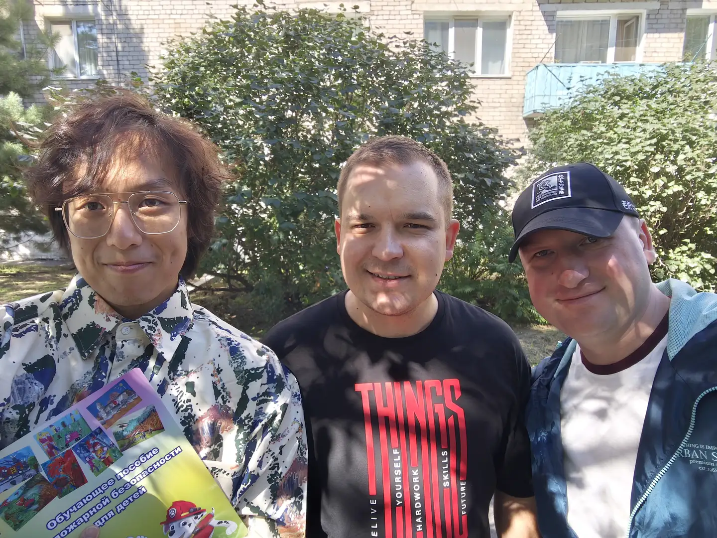 三位男士在户外合影，中间一人穿着黑色T恤，上面印有红色英文“THINGS”，左侧一人手持一本关于消防安全的儿童读物，右侧一人戴黑色棒球帽，穿着蓝色外套。背景是绿树和一栋浅色砖墙建筑，阳光明媚。