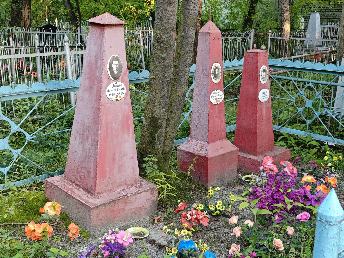 墓地中排列着三座粉红色的尖顶石碑，每座石碑上都镶嵌着圆形肖像和姓名信息。石碑周围种植着五颜六色的花朵，包括粉色、紫色、橙色和黄色。背景是铁艺围栏和树木，地面覆盖着绿色苔藓和落叶。