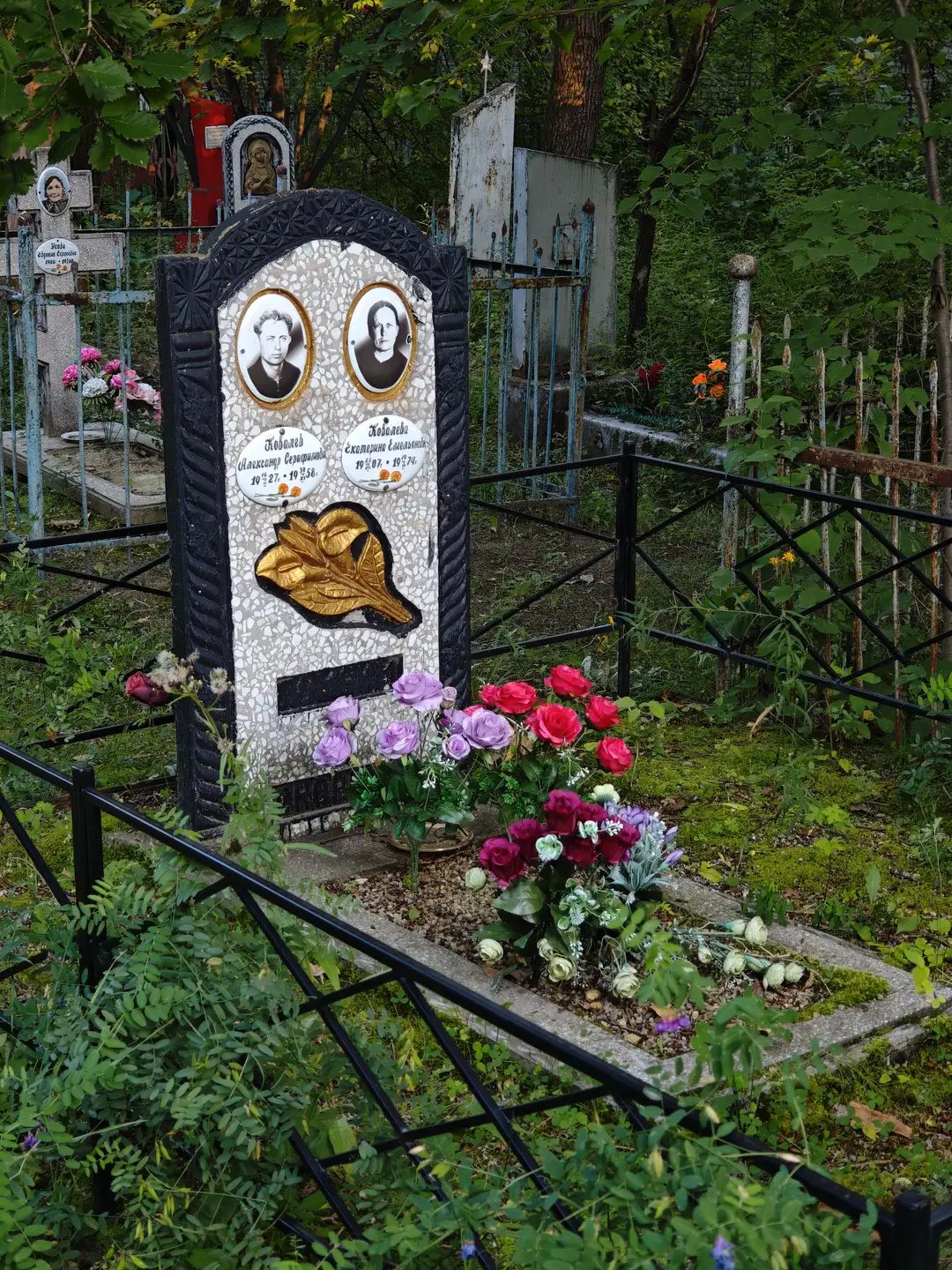 墓碑由黑色边框和白色石板构成，上面镶嵌着两幅人物肖像和名字信息，下方有一片金色叶子装饰。墓碑前摆放着红色、紫色和白色的鲜花，周围是黑色金属围栏。背景中可见其他墓地和绿色植被。