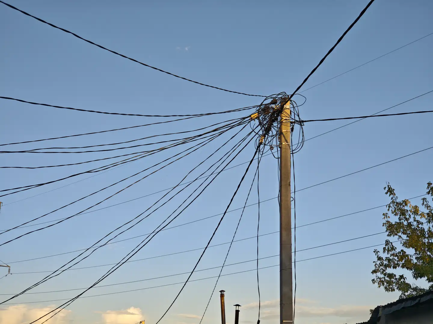 一根电线杆矗立在画面中央，顶部连接着多条纵横交错的电线。天空是清澈的蓝色，右下角有一棵部分可见的树。电线在空中形成复杂的网络，背景中还有几根烟囱。