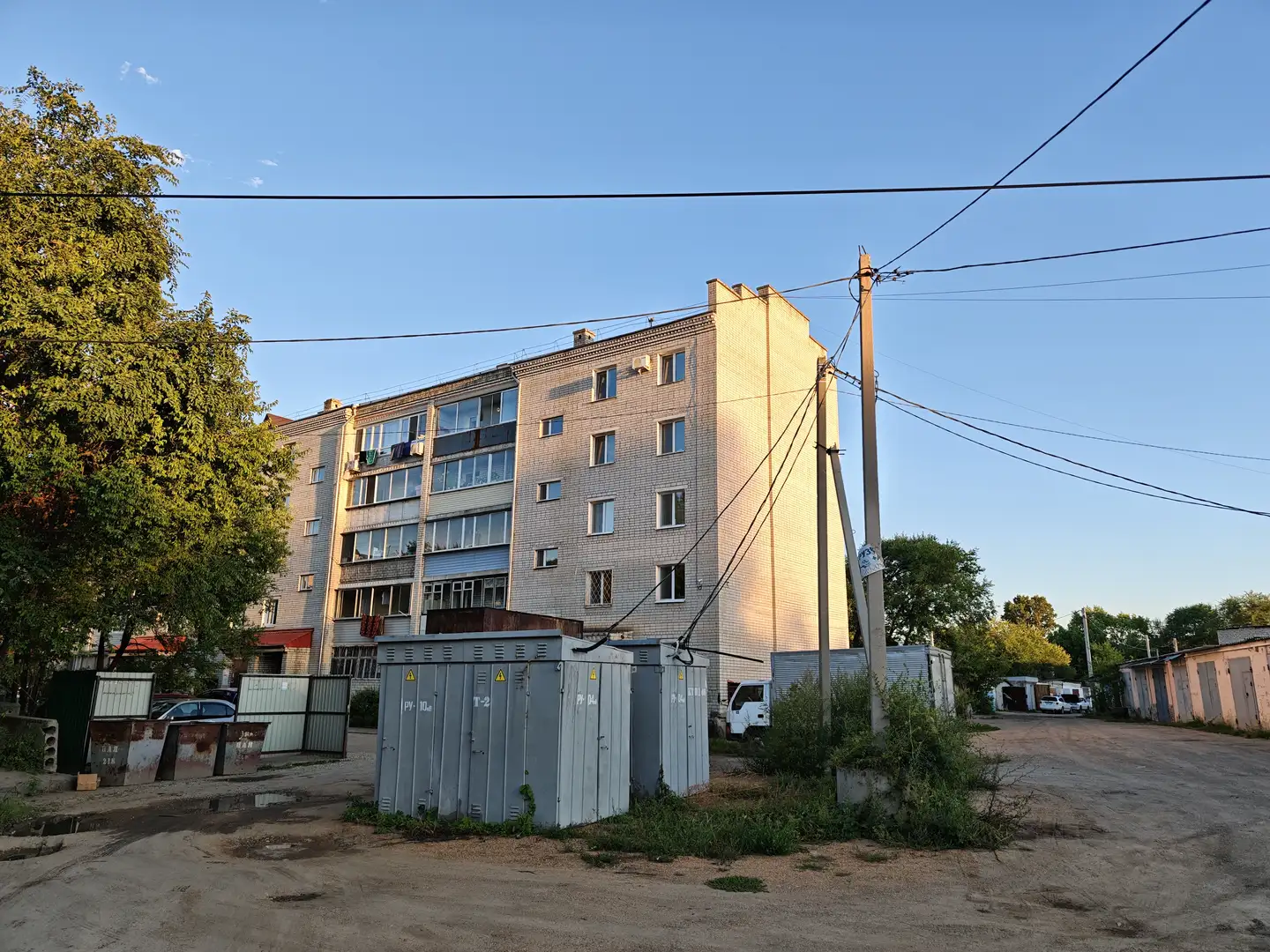 一栋五层高的住宅楼矗立在画面中央，外墙为浅色砖块，窗户排列整齐。楼前有几台灰色的电气箱和一些低矮的建筑。右侧有一根电线杆，天空晴朗，阳光洒在建筑物上。