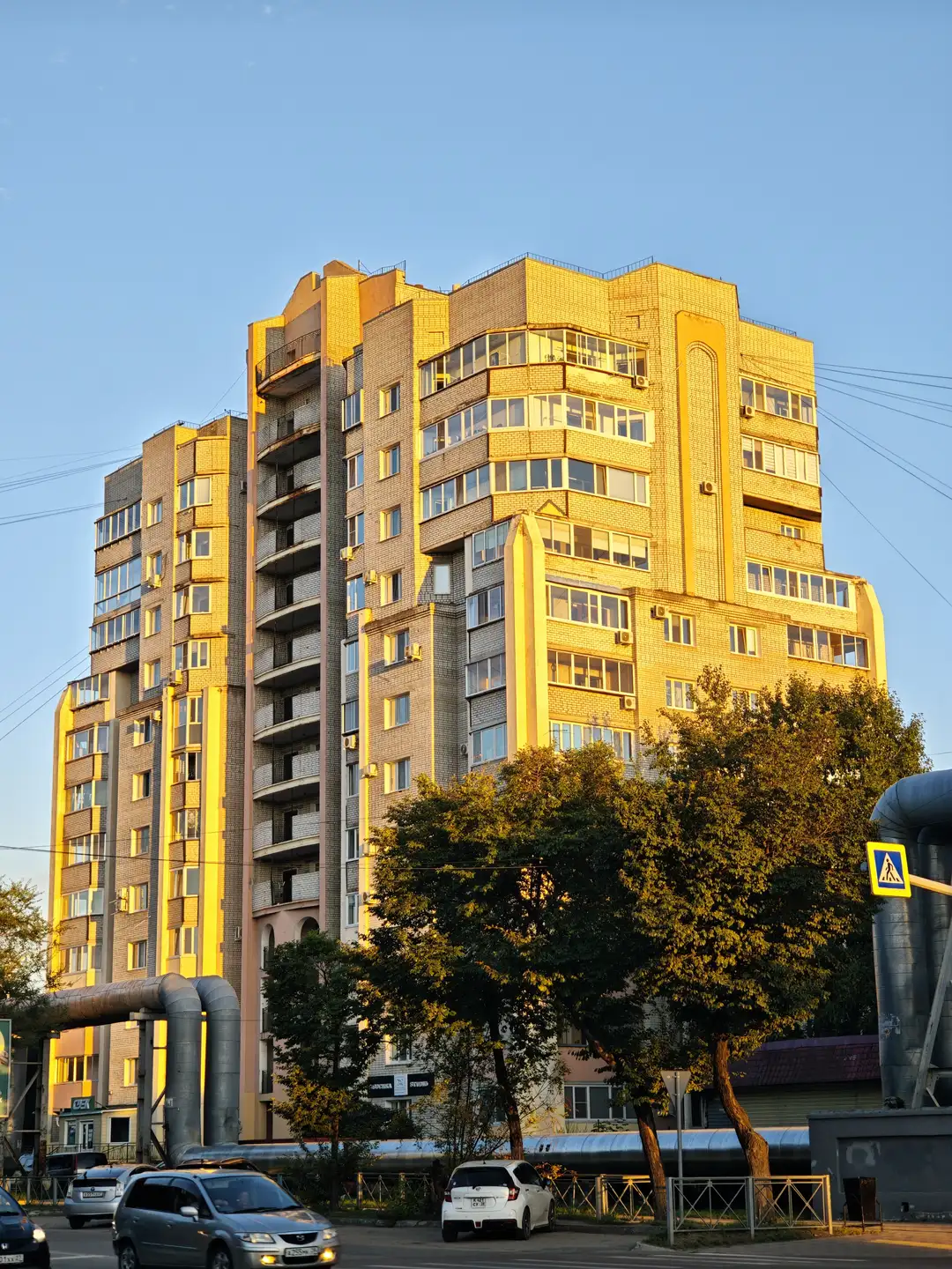 一栋高层住宅楼在夕阳下闪耀着金黄色的光芒，外立面由瓷砖和玻璃窗组成。楼体有多层阳台，窗户排列有序。前景中有几棵树和一辆行驶中的汽车，右侧有一个行人过街标志。