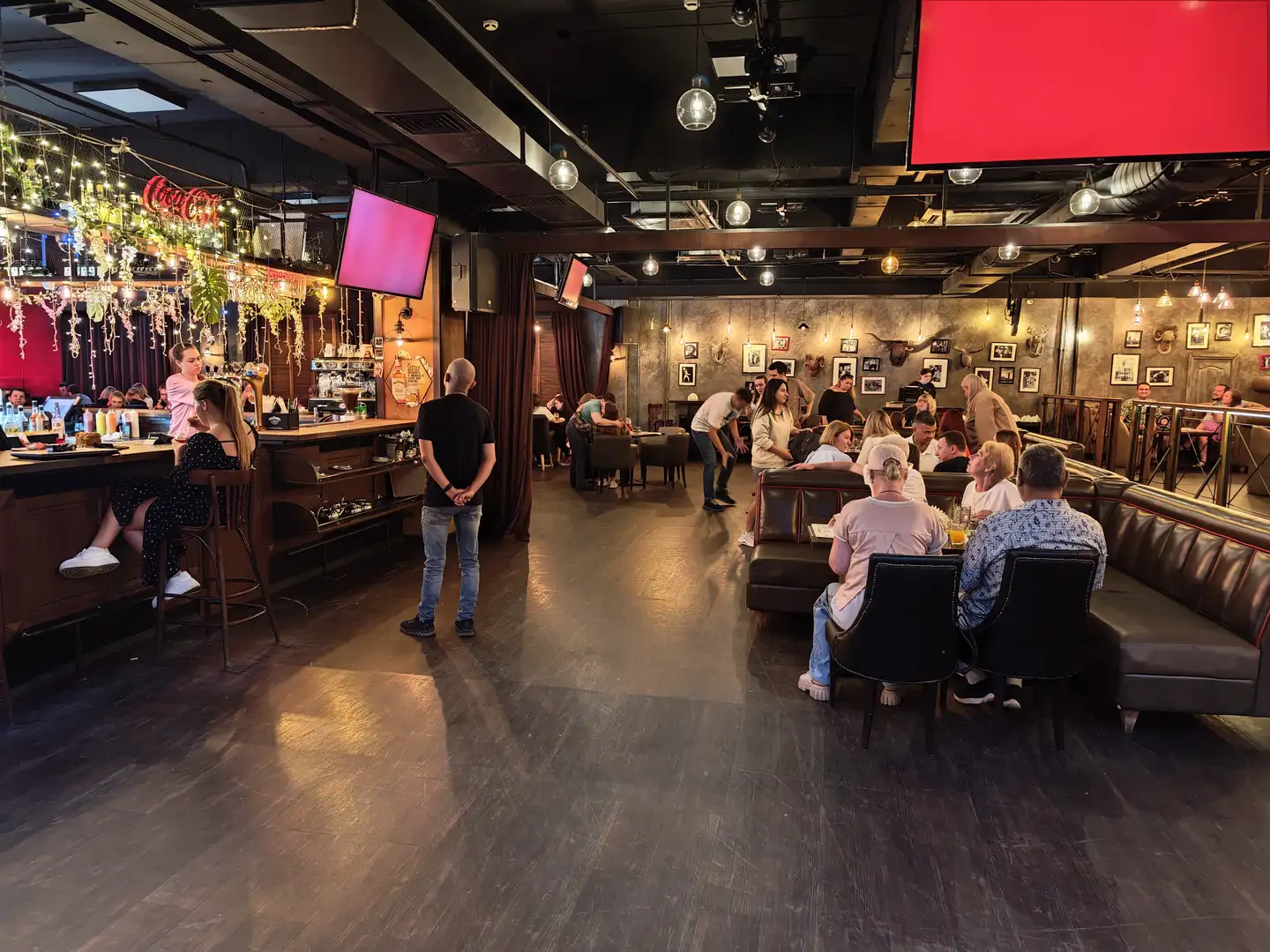 酒吧内部宽敞明亮，左侧是吧台，上方悬挂着装饰灯串和绿色植物。右侧有多个沙发座位，人们坐在那里交谈或用餐。天花板上安装了多个吊灯和屏幕，墙上挂着装饰画。