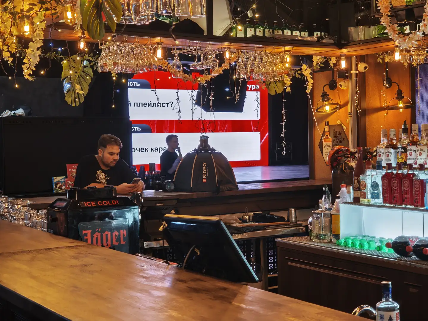 吧台后方有两名工作人员，一名正在看手机，另一名手持麦克风。吧台上摆放着酒瓶、杯子和调酒工具，上方悬挂着倒置的酒杯和装饰灯串。背景中有一个大屏幕显示文字内容。