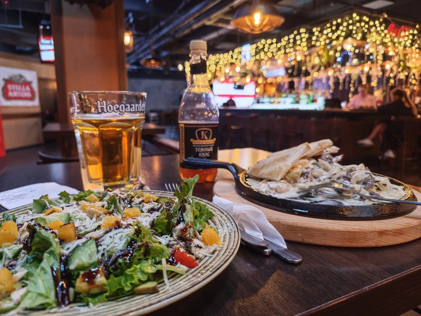 桌上摆放着一份沙拉和一份披萨，沙拉上有生菜、橙子块、奶酪和酱汁，披萨上覆盖着奶油酱和配料。旁边有一杯啤酒和一瓶饮料，背景中可以看到酒吧的灯光和装饰。