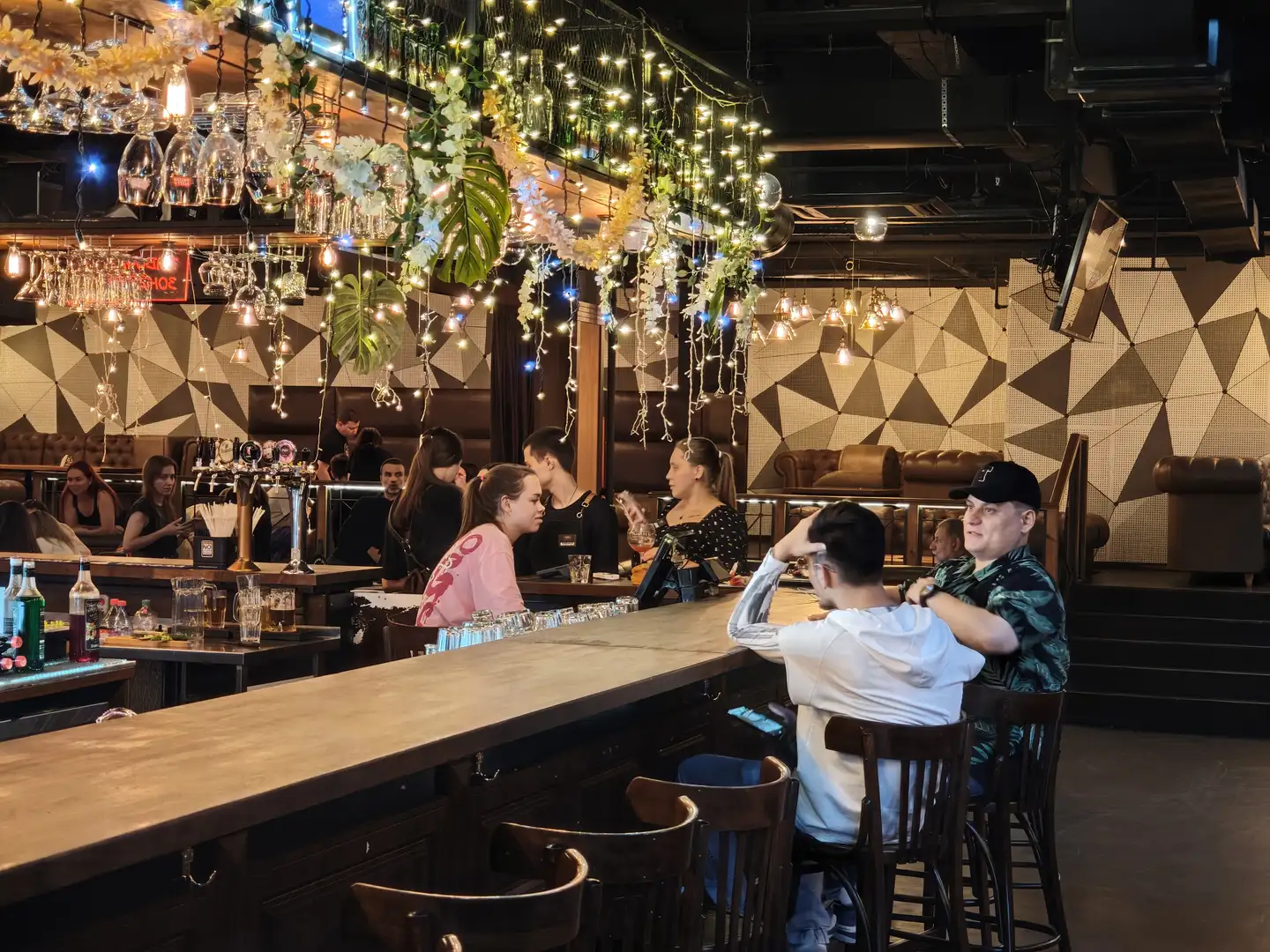 吧台上方悬挂着装饰花环和串灯，玻璃杯倒挂在架子上，几位顾客坐在吧台前与调酒师交谈
