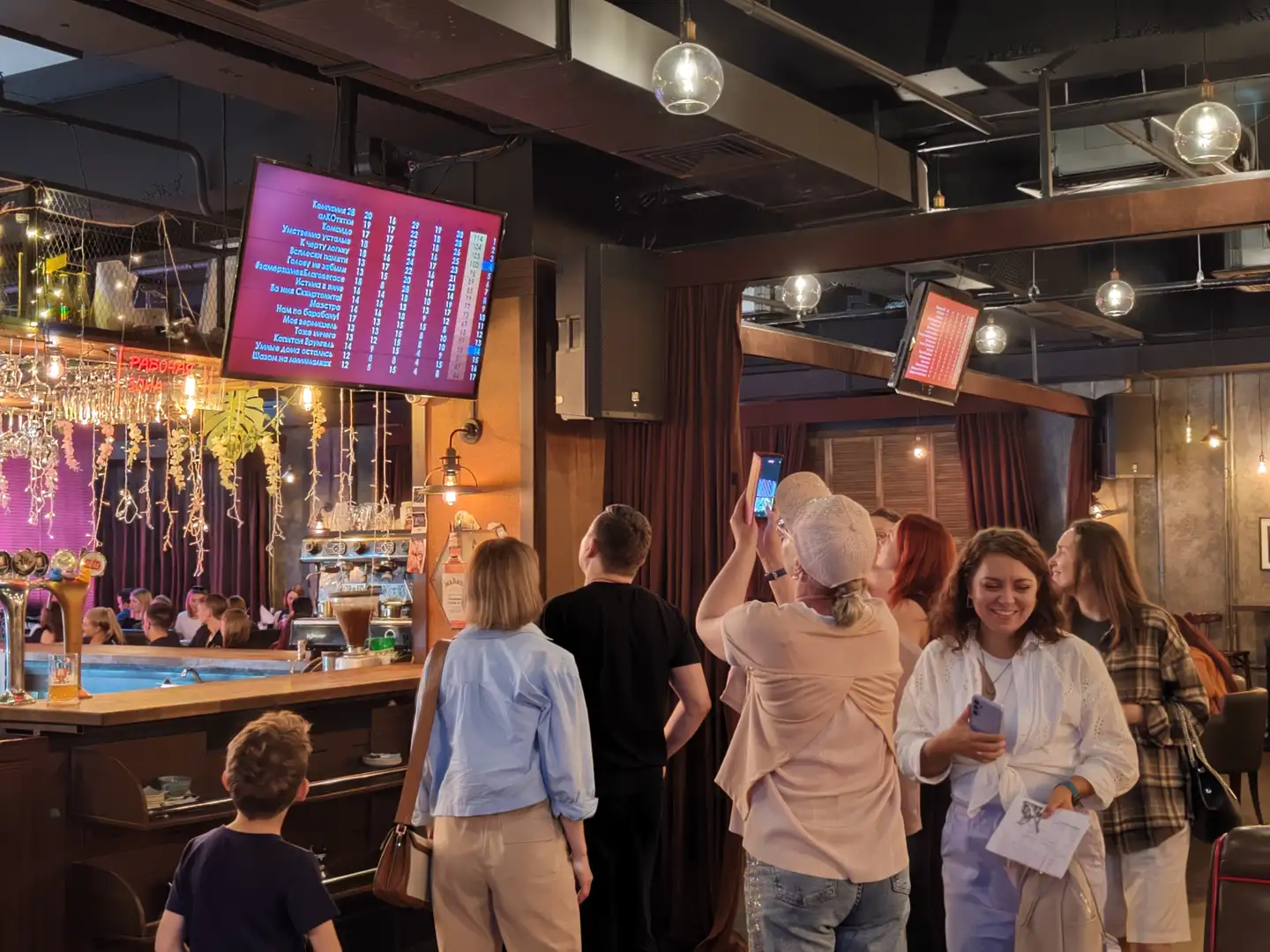 一群人站在酒吧区域，抬头看向墙上的电子屏幕，屏幕上显示着比赛得分和队伍名称，有人正在用手机拍照