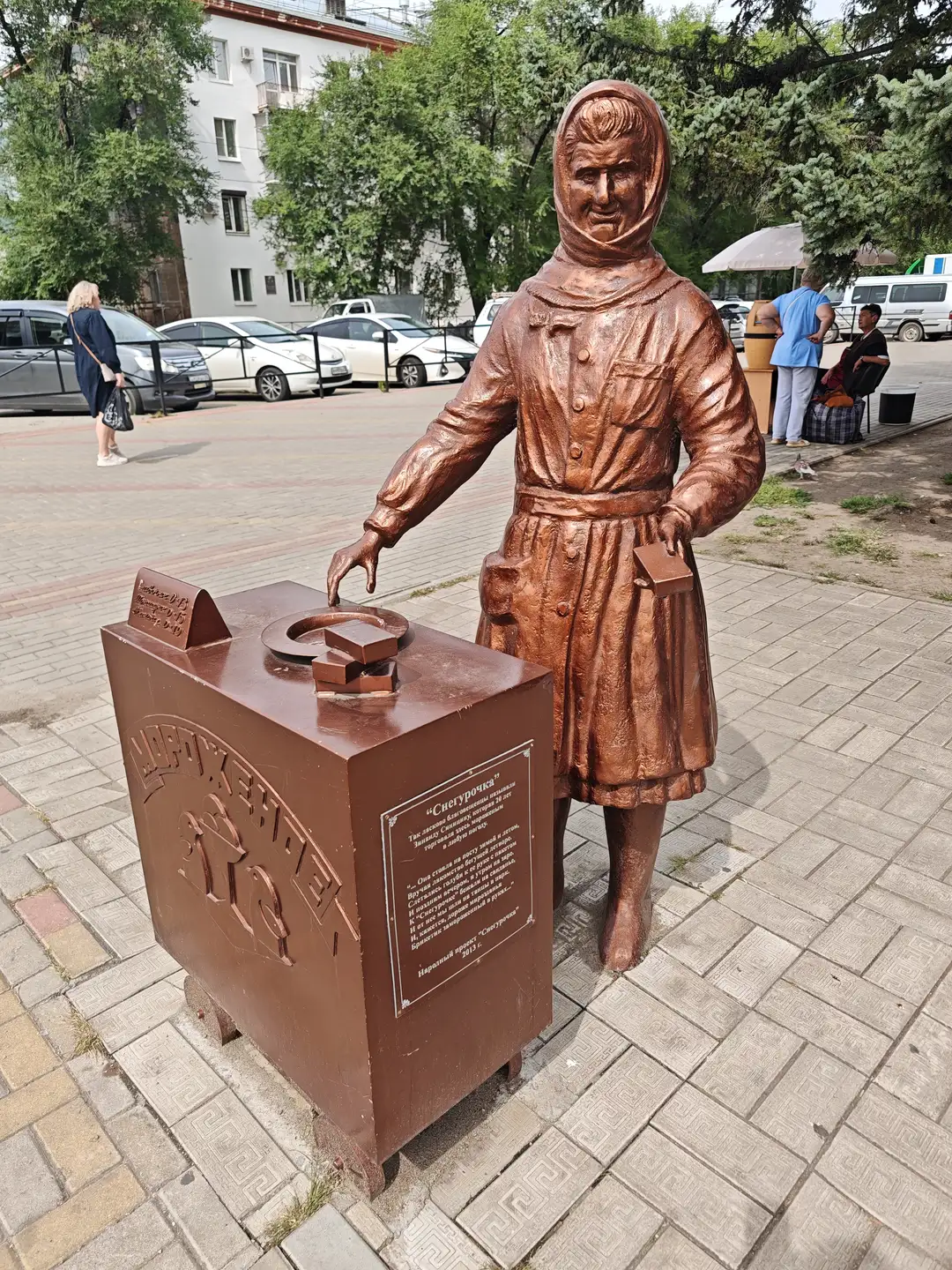 铜色的女性雕像站在一个类似收银台的物体旁，她身穿长裙和头巾，右手放在台上，左手拿着一块小牌子。收银台上有几块巧克力形状的装饰物，侧面刻有俄文文字和图案。背景是铺砖的广场，远处有行人、汽车和建筑物。