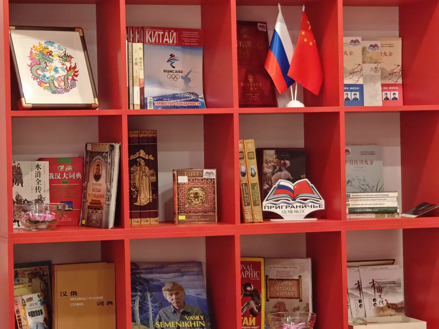 红色书架上陈列着各种书籍和装饰品，包括中文和俄文书籍、中国风画作、中俄两国国旗以及一个写有“ПРИГРАНИЧЬЕ 边境地区”的书立。书架上方还摆放着一本关于北京冬奥会的杂志和一些文化宣传册。整体布局整洁有序，突出中俄文化交流的主题。