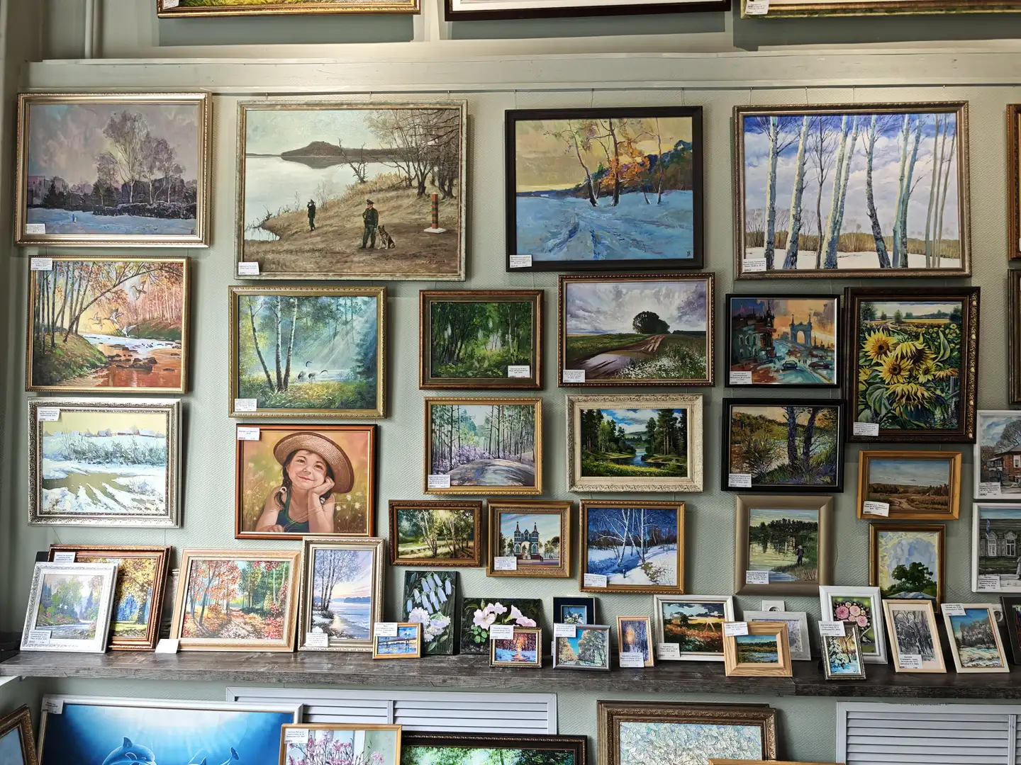 墙上挂满了各式各样的画作，以自然风景为主，包括森林、河流、雪地和乡村景色。画框大小不一，颜色多样，部分画作下方附有标签说明。画面中央有一幅人物肖像画，周围环绕着其他描绘季节变化与自然风光的作品。