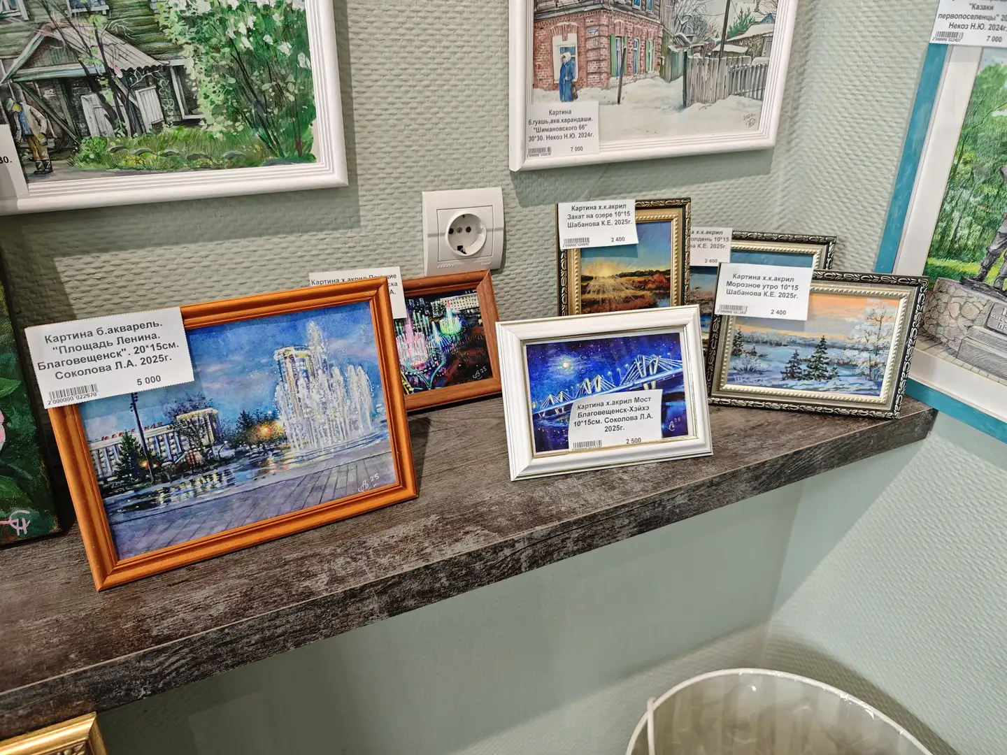 木质展台上摆放着几幅小型画作，画作内容为城市夜景和冬季景观，如喷泉广场和桥梁。每幅画前都贴有价格标签，标明了作品名称、尺寸、作者和年份。背景墙上也挂有多幅画作，墙面为浅绿色，带有纹理。