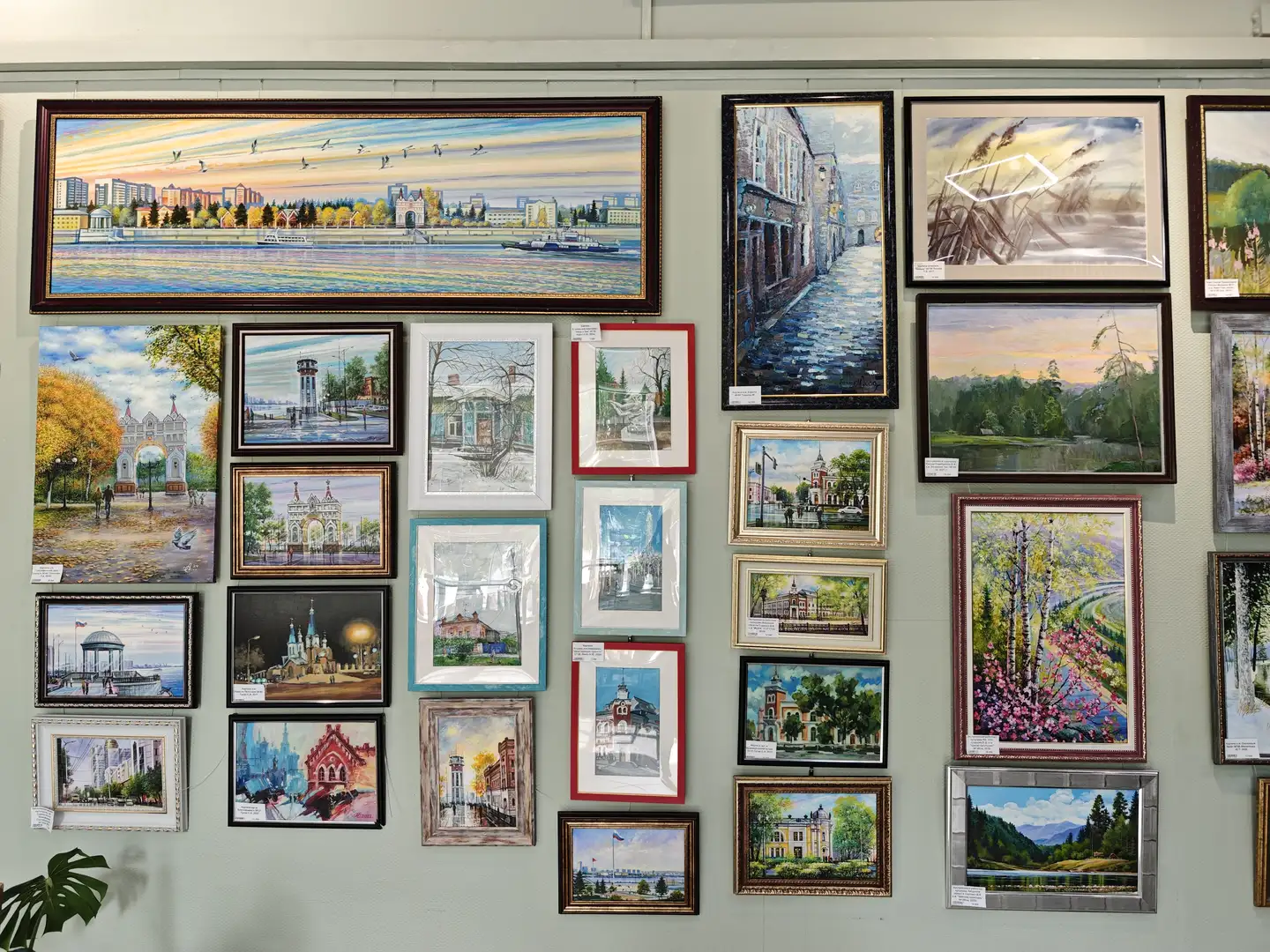 墙上挂满了不同尺寸和风格的画作，主题涵盖城市景观、建筑、自然风光和静物。画作排列紧密，色彩丰富，部分画作描绘了秋天的树木、河流和城市街道。整体布局呈现出艺术展览的氛围。