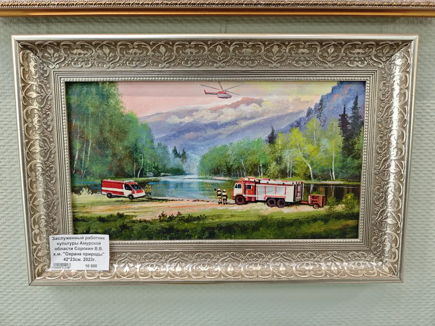 一幅装在银色雕花画框中的油画，描绘了一条河边的场景，有消防车、救护车和直升机，背景是山林和天空。画作下方的标签显示这是由阿穆尔州文化功勋工作者创作的，题为“自然保护”，尺寸为42×23厘米，创作于2023年。