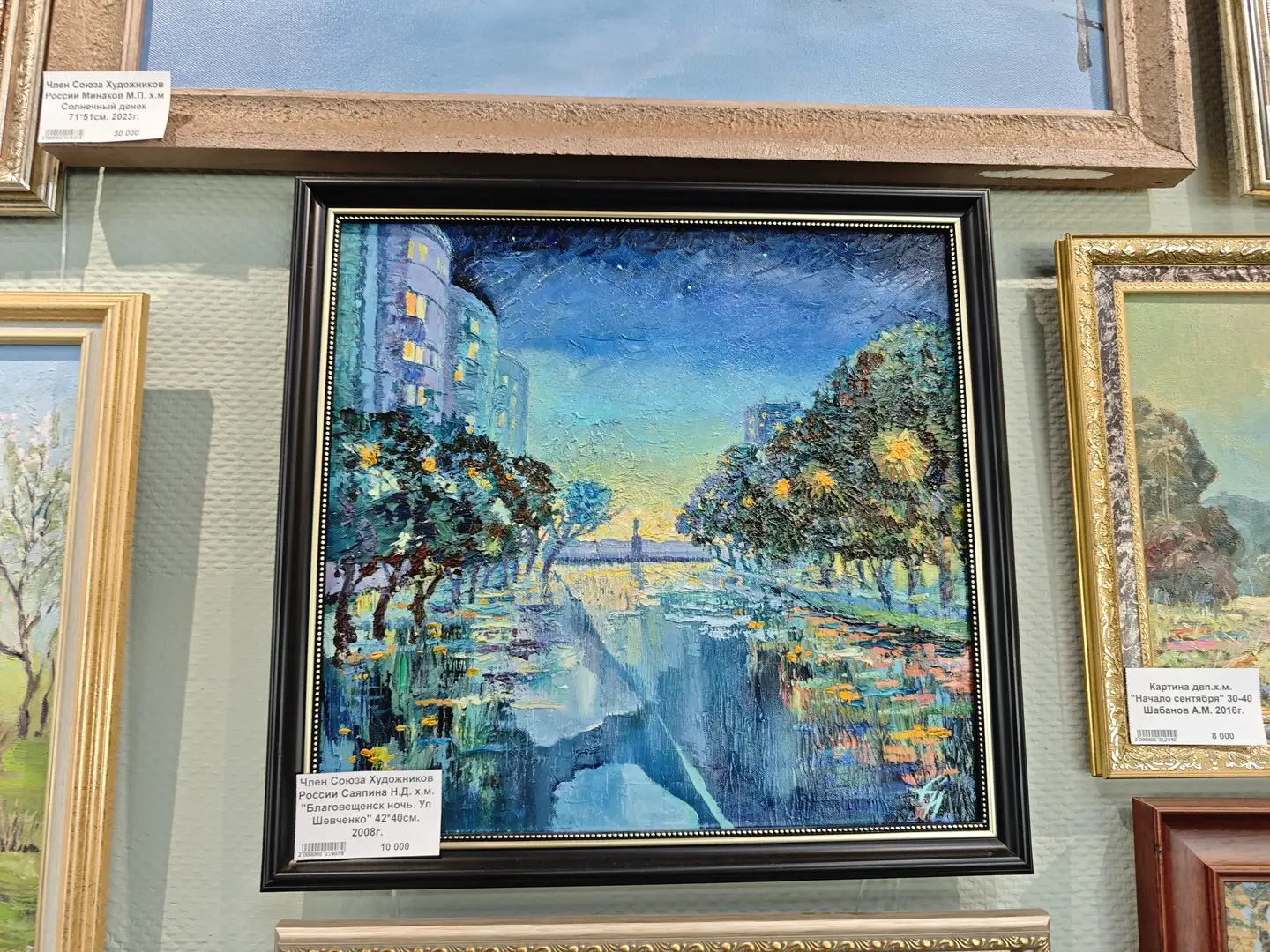 一幅黑色边框的油画，描绘了夜晚的城市河畔景色，河水倒映着两岸的灯光和树木，天空呈现深蓝色调。画作下方的标签注明作者为俄罗斯艺术家协会成员萨普林娜，作品名为“布拉戈维申斯克之夜”，尺寸为42×40厘米，创作于2008年。