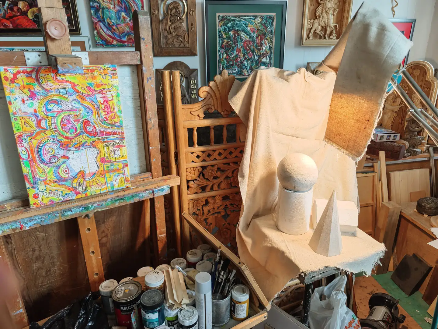 画室中有一个木制画架，上面放着一幅色彩鲜艳的抽象画，旁边是一个覆盖着布的模特台，上面摆放着几何形体。周围散落着颜料罐、画笔和其他绘画工具，墙上挂着多幅已完成的作品，包括宗教题材和抽象画。