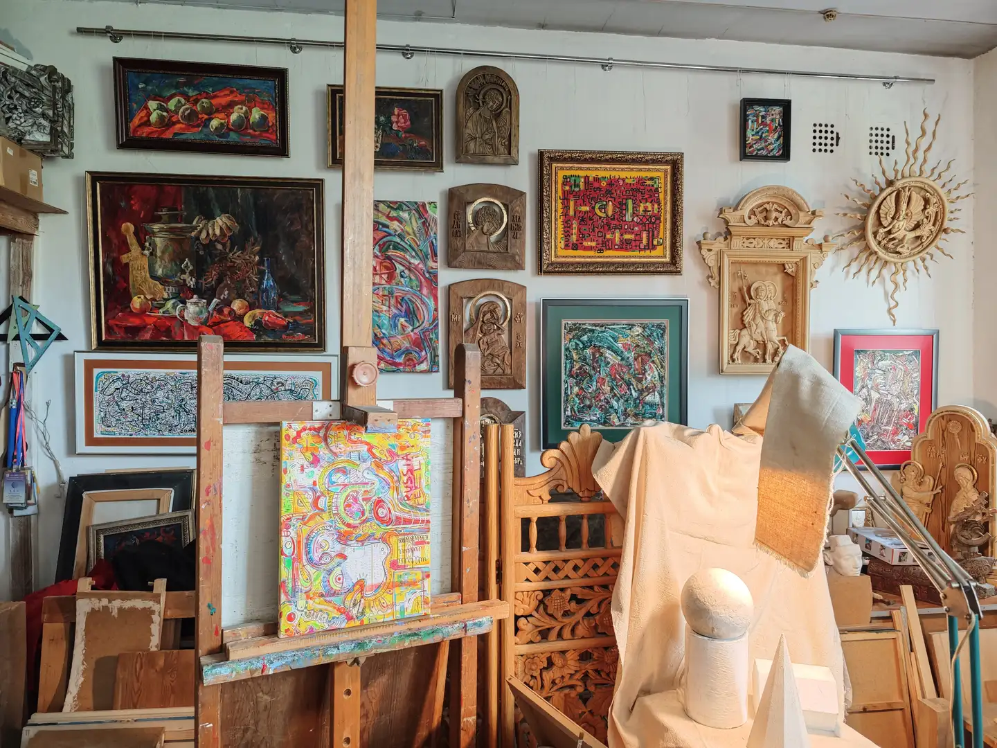 画室墙壁上挂满了各种艺术作品，包括静物画、宗教浮雕、抽象画和装饰性木雕。房间中央有一个画架，上面放置着一幅色彩丰富的抽象画，旁边是一个覆盖布的模特台，上面摆放着几何形体。地面和角落堆放着画框和木制品，环境显得充实而富有创作气息。