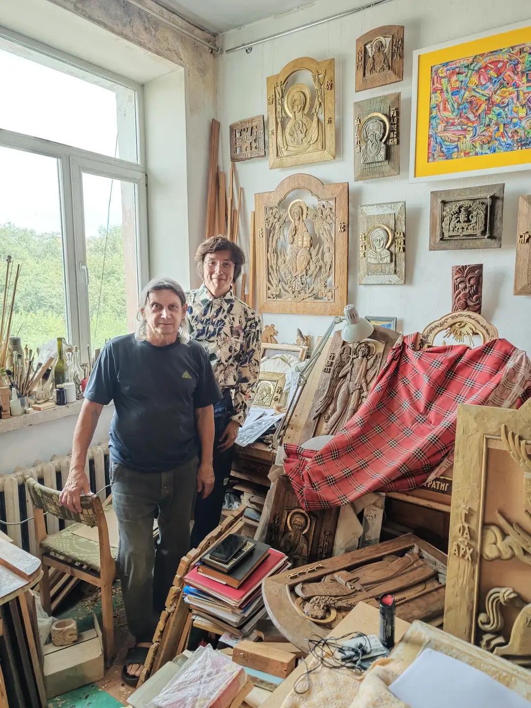 两位成年人站在一个充满木雕艺术品的工作室里，墙上挂满了宗教主题的木雕作品。房间内有雕刻工具、木板和一些书籍，窗边摆放着各种材料和用品。一位穿着深色T恤，另一位穿着花纹衬衫，两人面带微笑。