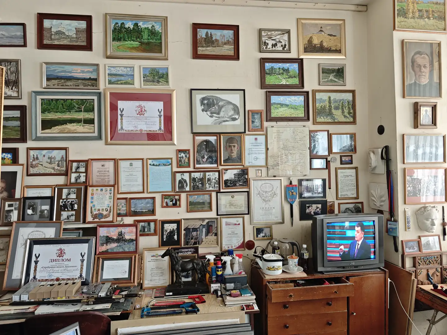 墙上挂满了各式各样的画作和证书，包括风景画、人物肖像和动物素描。下方的桌面上摆放着文具、书籍、一个黑色马雕像以及一台正在播放节目的老式电视机。房间显得非常拥挤但充满艺术气息。