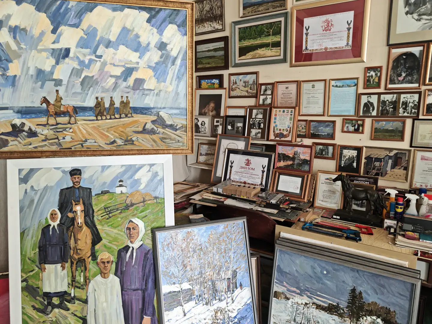 墙面上挂满了多幅画作和奖状，其中包括一幅描绘海边骑马场景的大画，以及一幅乡村人物群像。桌面上堆叠着未装裱的画作，内容为雪景和自然风光。整个空间布满艺术作品，展示出浓厚的创作氛围。