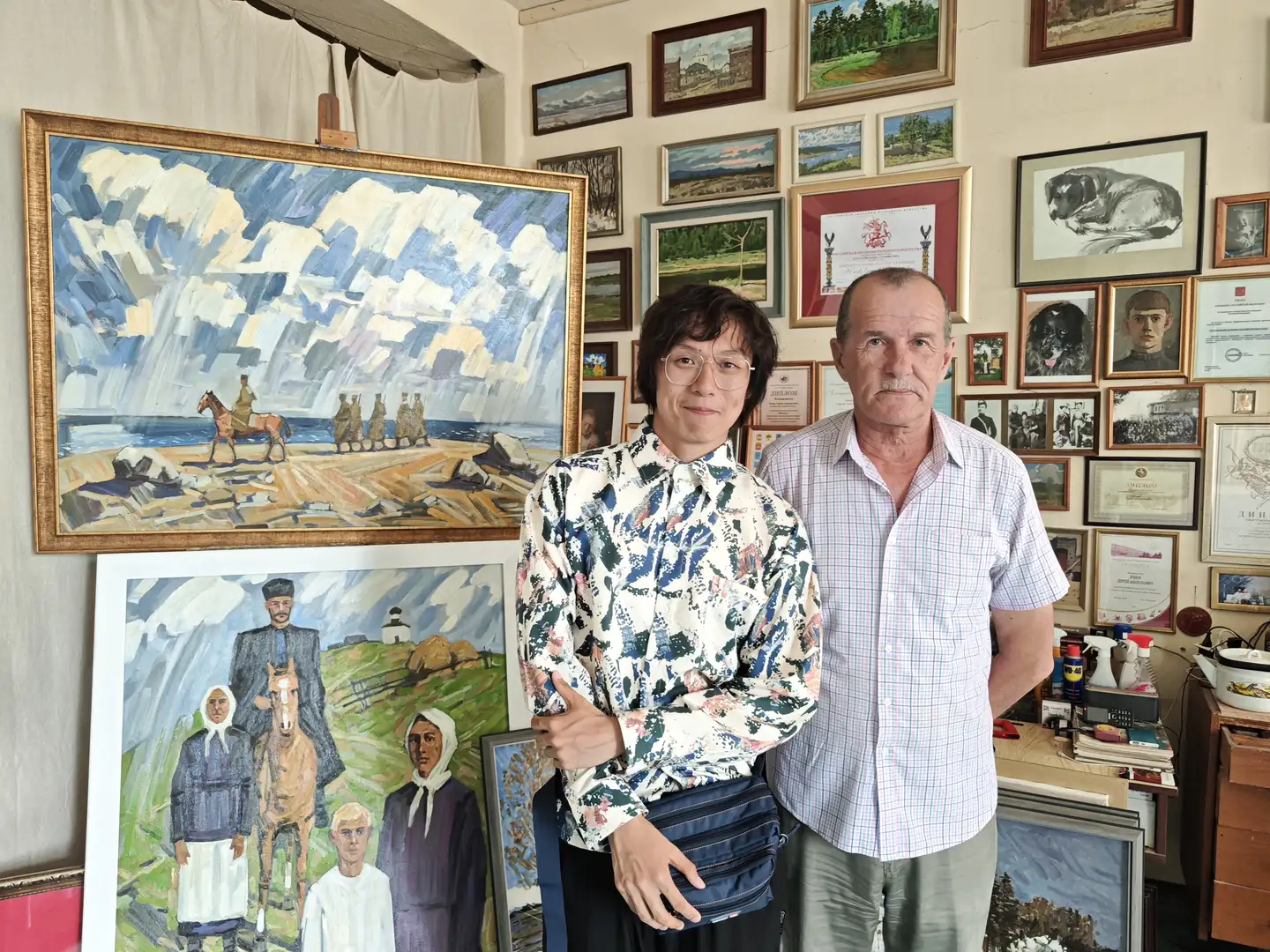 两位男士站在一间装饰满画作和证书的房间里，其中一人穿着花衬衫，另一人穿着格子衬衫。背景中有一幅大型海景画和一幅乡村人物画，墙上挂满了各种尺寸的画框和奖状。房间内陈设整齐，充满艺术气息。