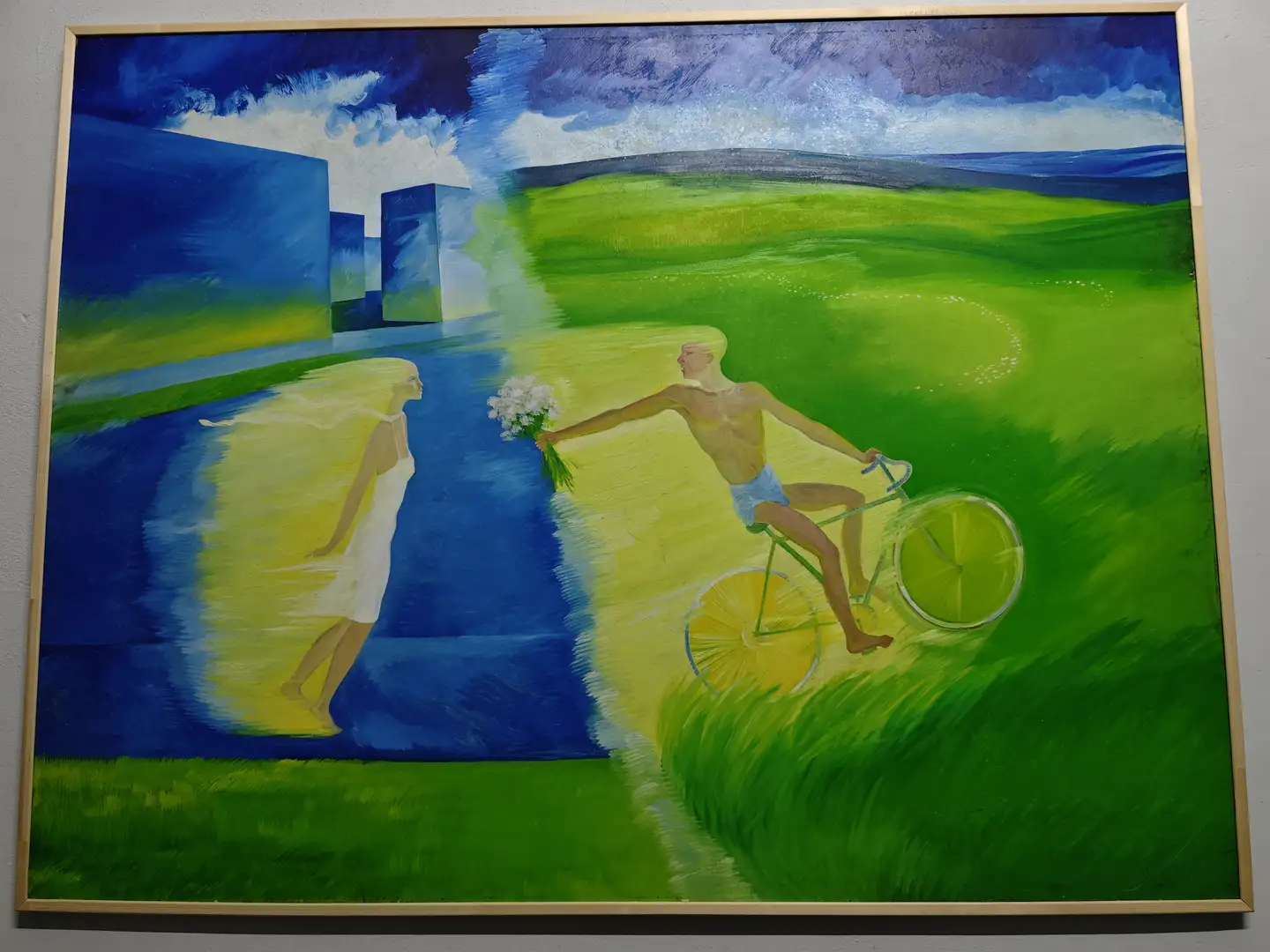 画作展现了一位骑自行车的男子在绿色草地上飞驰，手中拿着一束白色花朵。画面左侧是一位身穿白色长裙的女子，身影透明，仿佛在水中倒影。背景是蓝色的几何建筑和广阔的天空，色彩鲜明且富有动感。