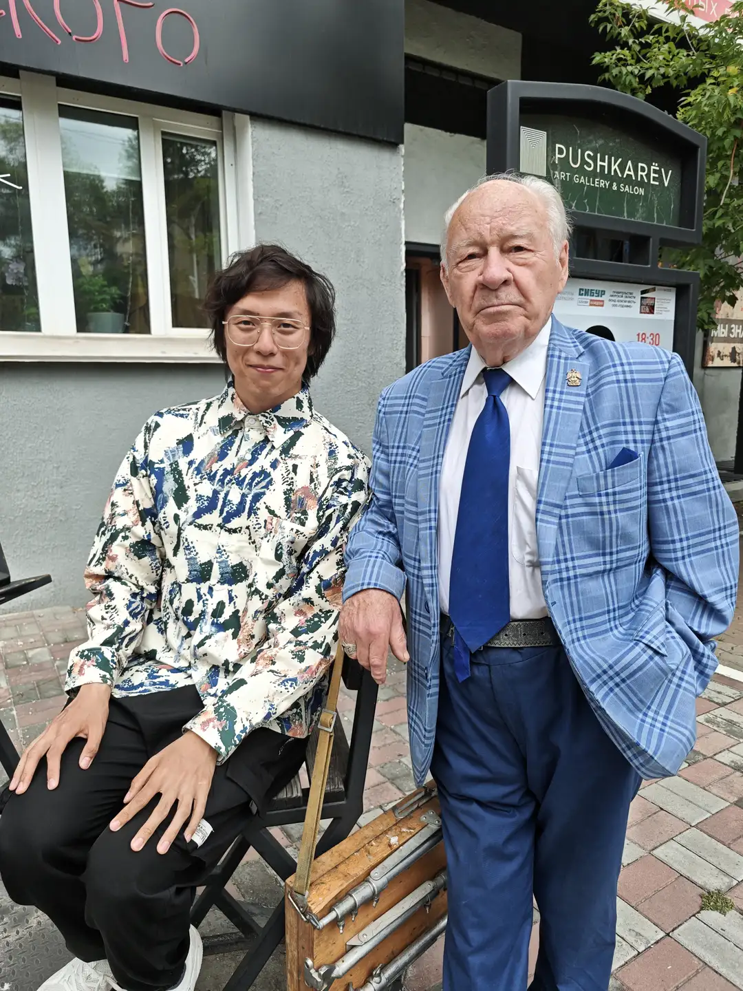 一位年轻男子坐在椅子上，旁边站着一位年长的男士，两人站在一家名为“PUSHKAREV ART GALLERY & SALON”的画廊前。年轻男子穿着印花衬衫和黑色裤子，年长者穿着蓝白格纹西装外套和蓝色领带。背景中可见绿植和部分建筑细节。