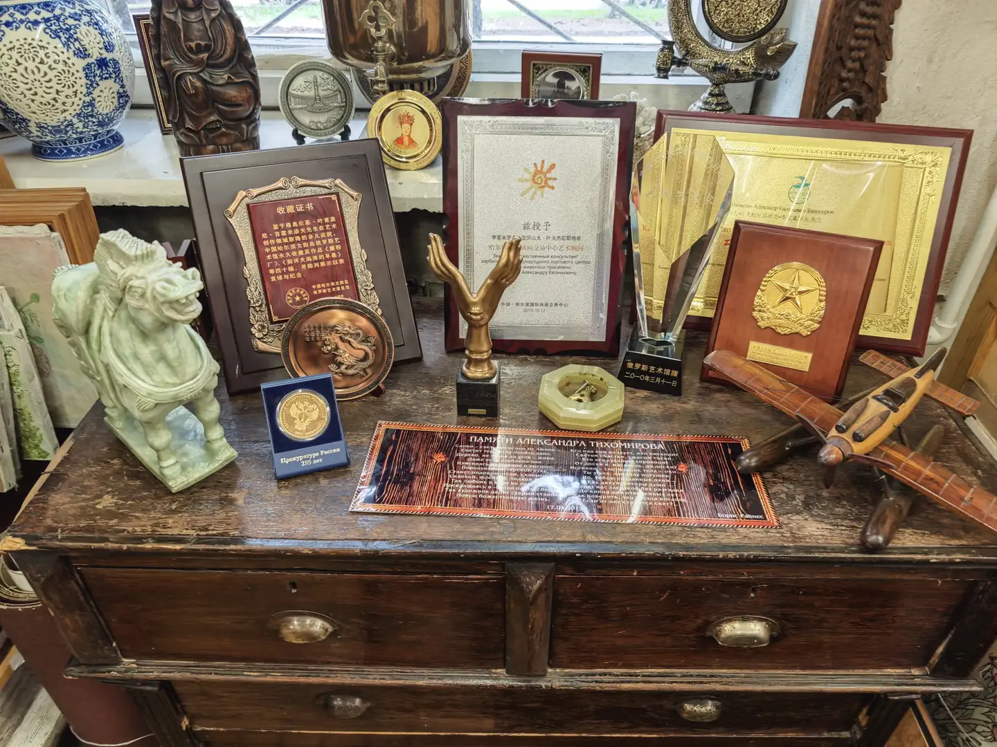 一张古旧的木桌上摆放着多个奖牌、证书和纪念品，包括一块红色底金色文字的证书、一个金色手掌雕塑、一枚圆形徽章和一架木质飞机模型。桌面上还有一块刻有俄文的金属牌匾，旁边是一尊青瓷石狮子和一个蓝白相间的瓷瓶。