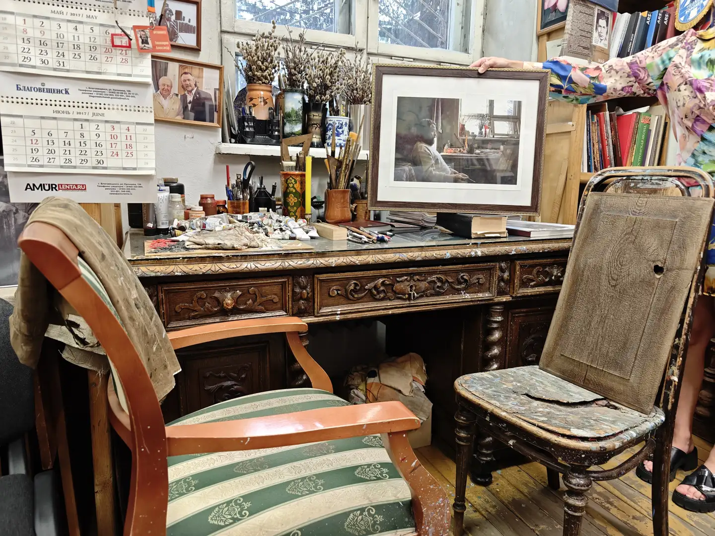 一张雕花木制书桌摆放在房间中央，桌上放着颜料、画笔和其他绘画工具，以及一幅装裱好的黑白照片。桌子旁有两把椅子，一把是条纹软垫椅，另一把是破旧的木椅。墙上挂着日历和几张照片，窗台上摆放着干花和陶器。