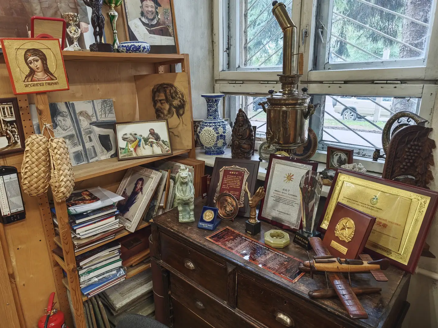 一个木制柜子上陈列着多种物品，包括奖状、一个铜制茶炊、一个蓝白瓷瓶和一个小型佛像。柜子上方的架子上放着书籍、画作和编织篮子，旁边是一扇窗户，窗外可以看到街道和车辆。柜子上还有一个木质飞机模型和一个绿色石雕小兽。