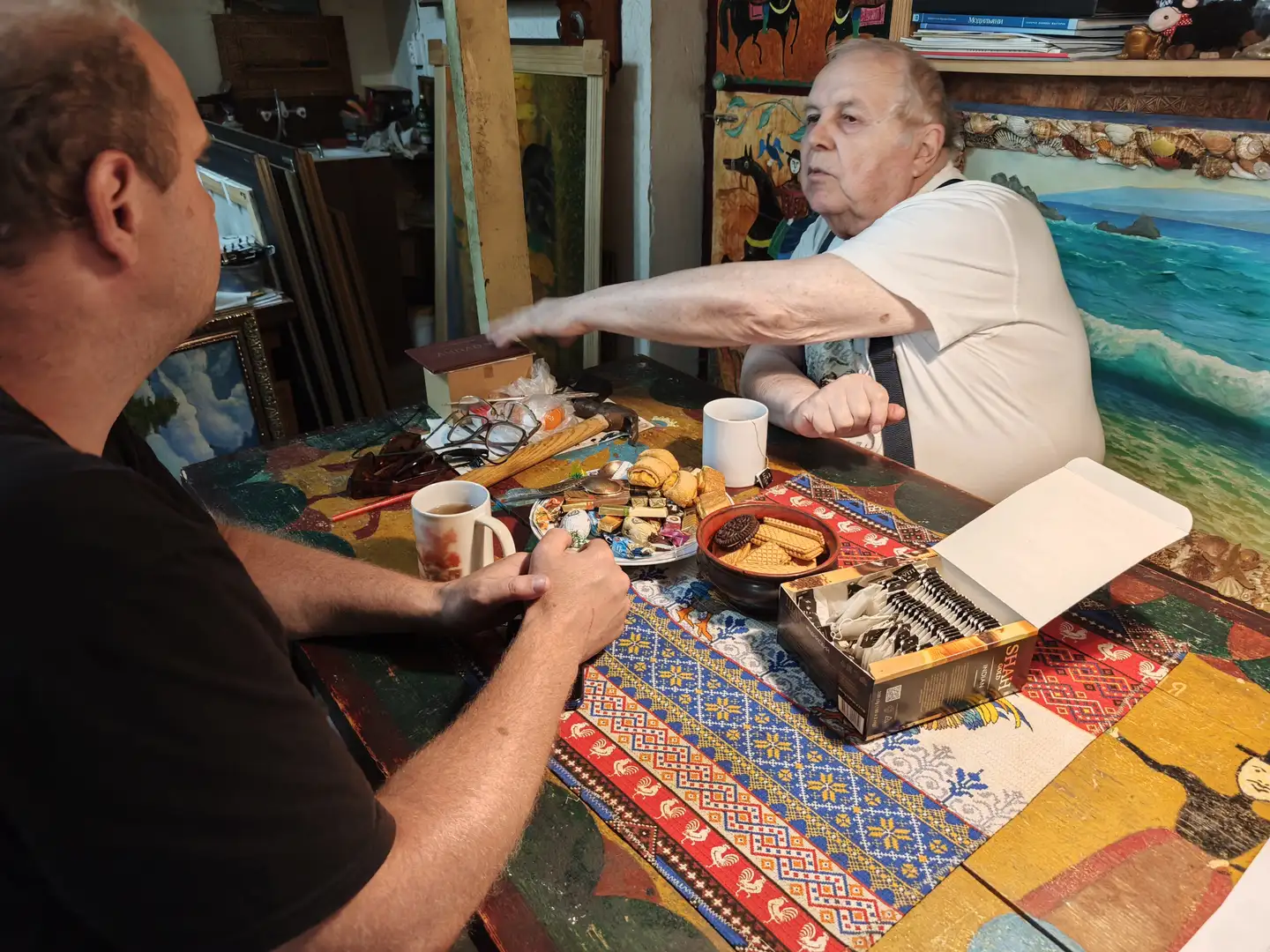 两位男士坐在一张装饰有彩色图案的桌子旁，桌上摆放着茶杯、点心和一盒象棋。一位年长的男士正伸手去拿桌上的物品，另一位则握着茶杯。背景中可见多幅画作和艺术用品，营造出一个充满艺术气息的环境。