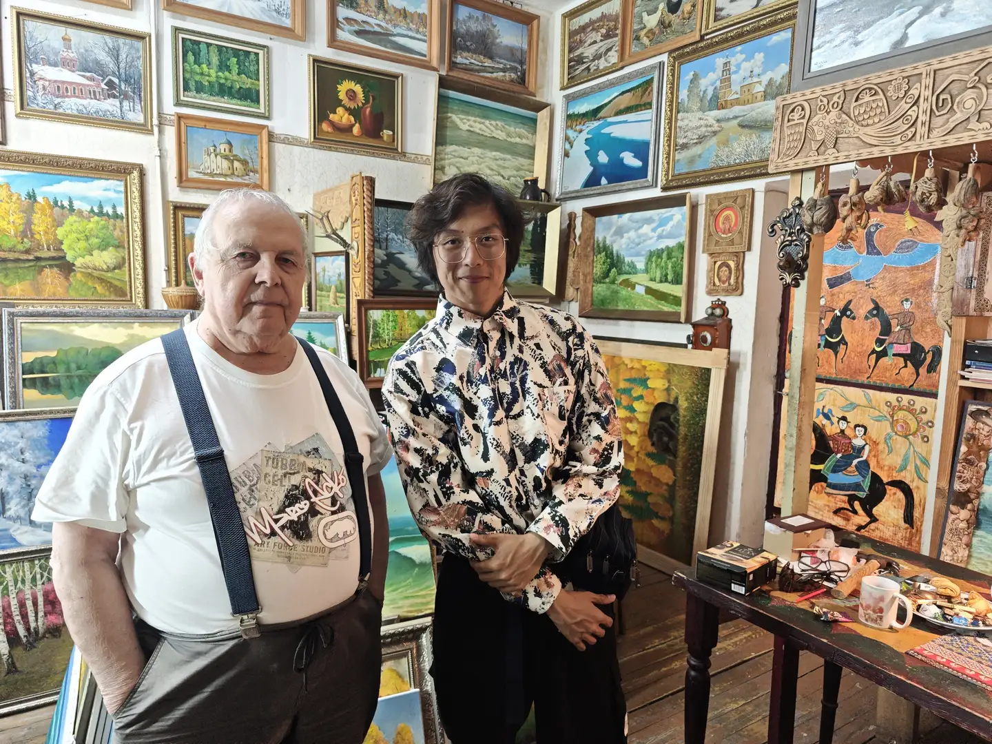 一位年长的男士和一位年轻男子站在一间挂满风景画的房间里，墙上陈列着许多装裱好的画作。年长者穿着白色T恤和背带裤，年轻男子穿着带有复杂图案的衬衫和黑色裤子。房间的一角有一张桌子，上面放着一些物品，包括一个杯子和食物。