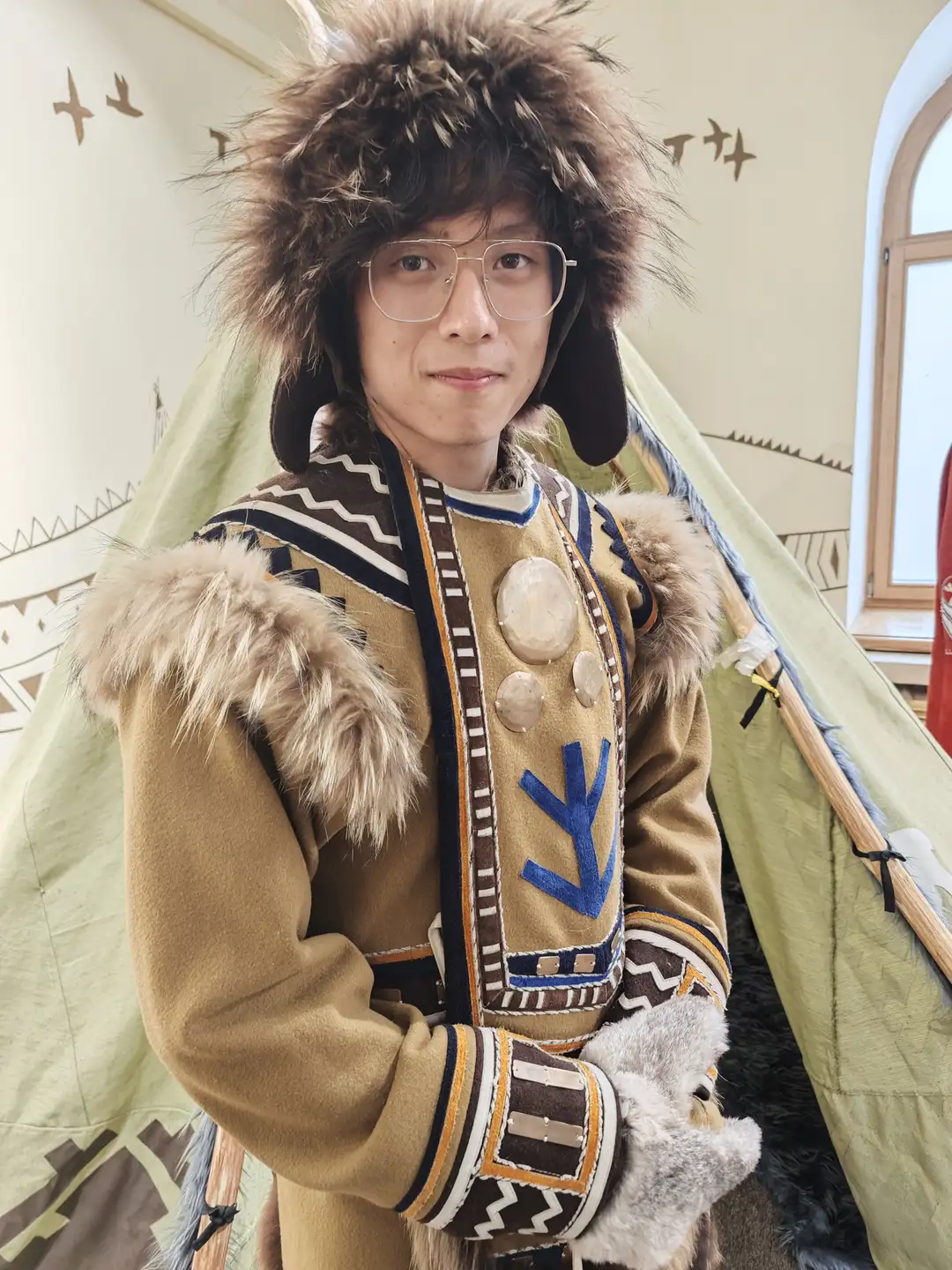 一位戴眼镜的年轻人穿着传统的毛皮边饰外套和毛帽，站在一个印有几何图案的帐篷前。他的外套上有蓝色箭头图案和圆形装饰，双手戴着毛茸茸的手套，面带微笑。