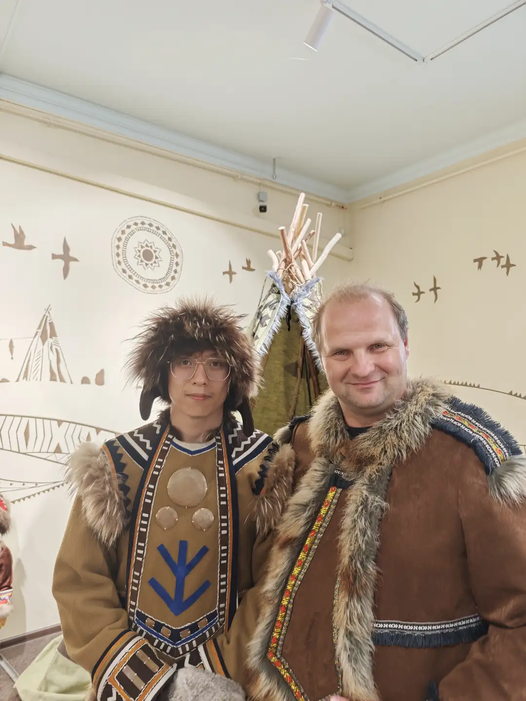 两位男士并肩站立，左边的年轻男子穿着带有毛皮边饰的传统服饰，右边的中年男子也穿着类似的毛皮外套。他们身后是一个装饰有飞鸟和帐篷图案的墙面，中间有一个小帐篷模型。