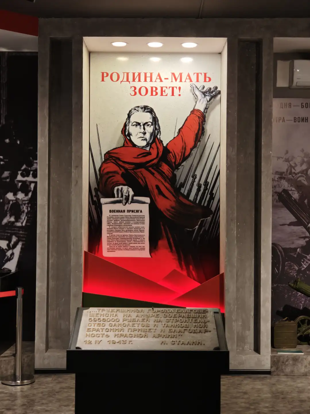 一幅大型宣传海报上写着“РОДИНА-МАТЬ ЗОВЕТ!”，描绘了一位身穿红色斗篷的女性，右手高举，左手拿着一份文件，背景是整齐排列的步枪。海报下方有一块石碑，刻有俄文文字和日期“12 IV 1943 г.”。