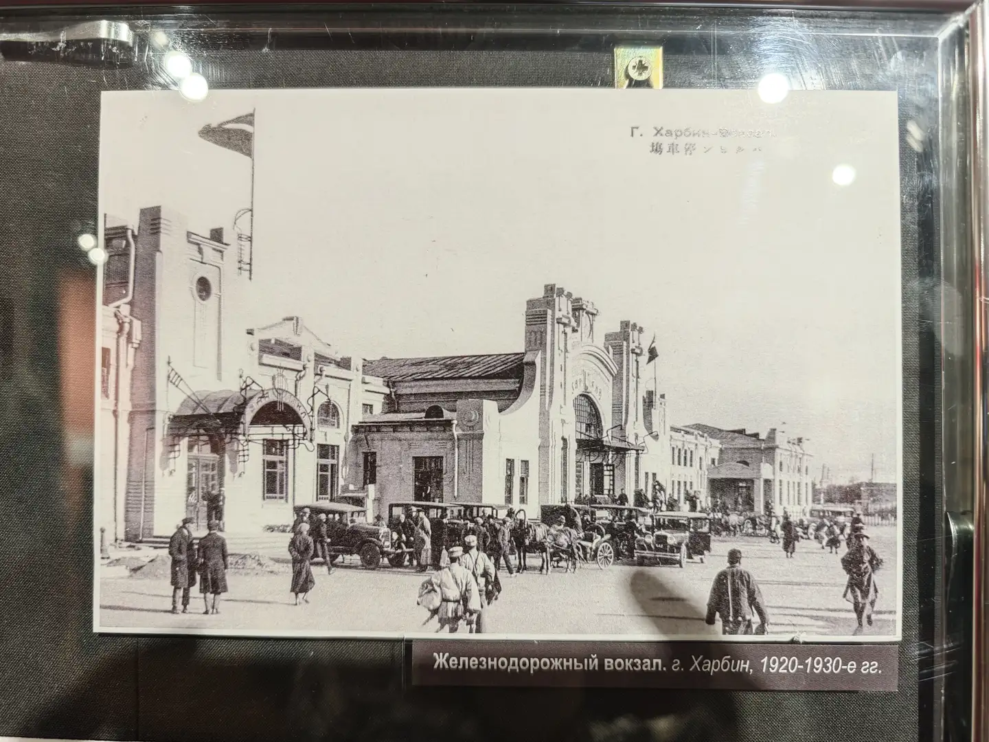 哈尔滨火车站前广场，建筑具有典型的苏联风格，门口聚集了许多人和车辆。画面中有马车、汽车和行人，显示出当时交通方式的多样性。照片下方标注为“Железнодорожный вокзал. г. Харбин, 1920-1930-е гг.”