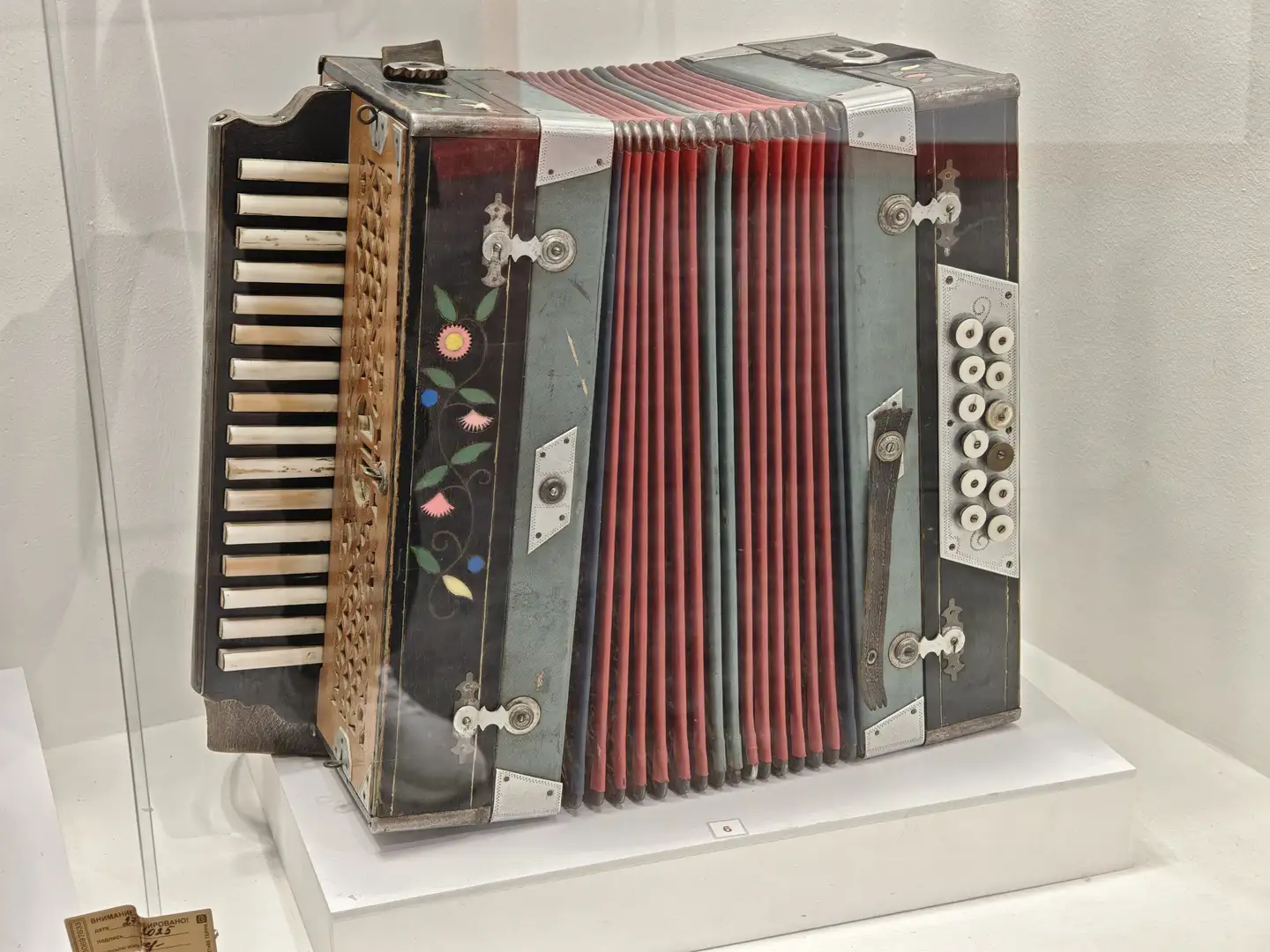 一把老式手风琴陈列在展台上，琴身由金属和木质材料构成，表面有花卉图案装饰。左侧是键盘部分，右侧是按钮，中间是红色风箱，整体略显陈旧但保存完好。琴身上可见磨损痕迹，表明其年代久远且曾被频繁使用。