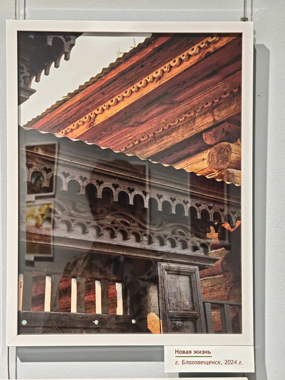 一幅展示传统木结构建筑局部的照片，屋顶由红色瓦片覆盖，屋檐下有精美的雕刻装饰。建筑的木质梁柱清晰可见，门框上方有拱形雕花，整体风格古朴。照片下方标注为“Новая жизнь г. Благовещенск, 2024 г.”