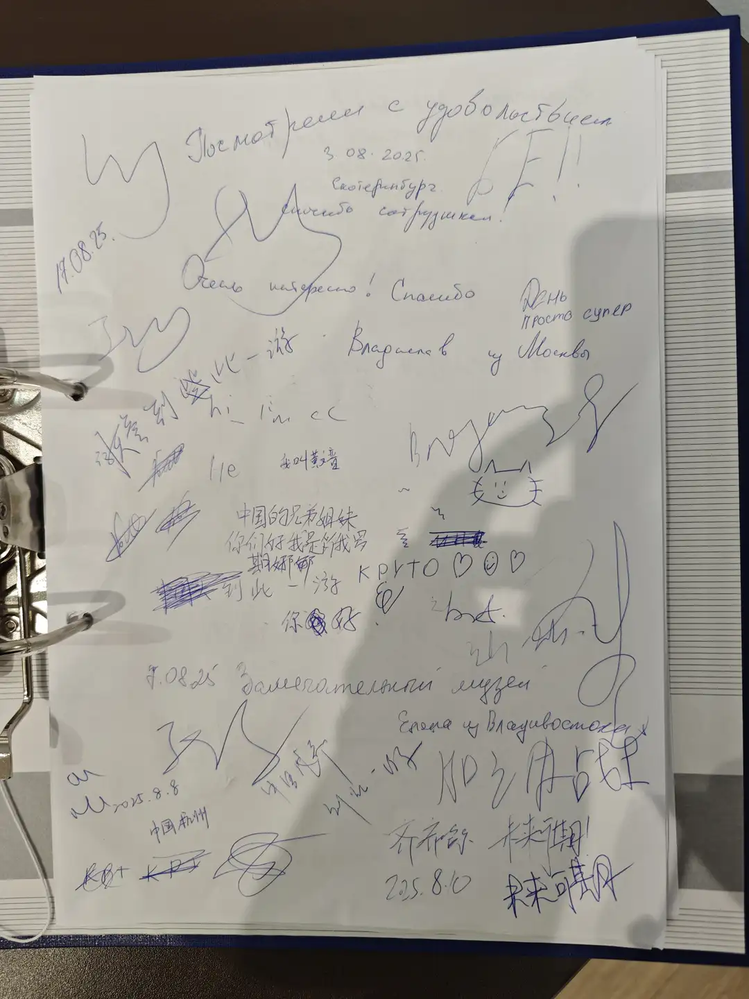 一本留言簿的一页，上面写满了不同语言的签名和留言，包括俄文、中文和英文。内容表达了对展览的兴趣和感谢，有的还画了笑脸或心形符号。日期显示为2025年8月，来自中国杭州、莫斯科等地的游客留下印记。