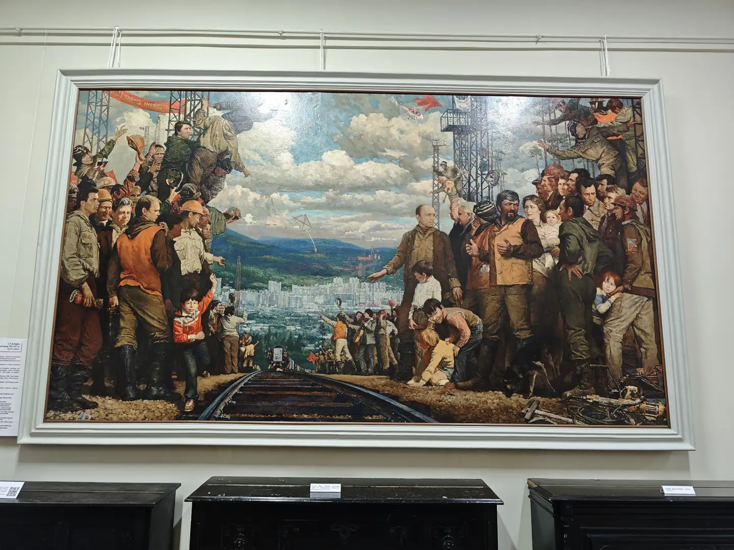 一幅大型油画，描绘了一群人在铁路轨道旁聚集，背景是正在建设中的城市。画面中人物穿着工装，有的举着红旗，有的抱着孩子，表现出劳动人民的团结与希望。远处山峦起伏，天空中有风筝飞舞，象征着新生活的开始。