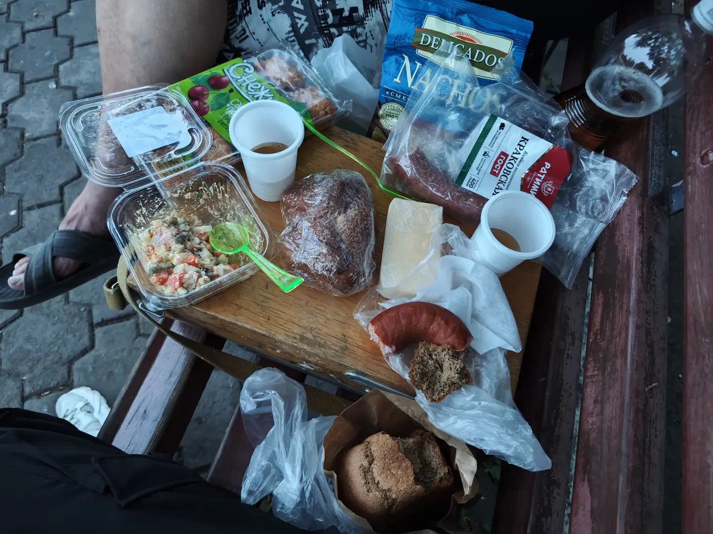 一张户外野餐桌上摆放着各种食物，包括面包、香肠、奶酪、沙拉和饮料。桌上有塑料盒、纸杯和包装袋，食物部分已被食用。旁边有人坐着，脚穿拖鞋，环境显得随意而放松。